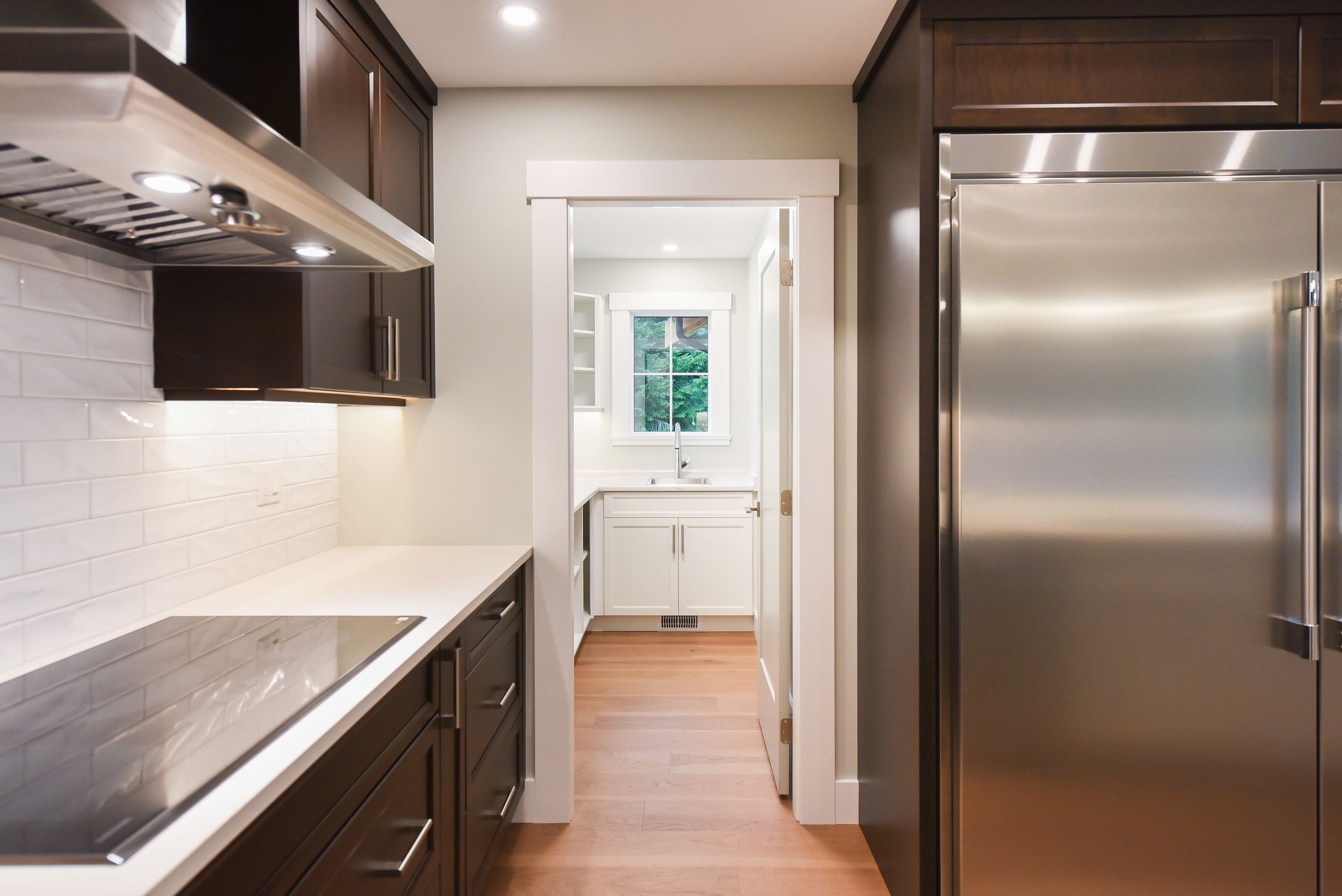 View of a modern kitchen with dark cabinets, a white countertop, stainless steel appliances, and a doorway leading to a bright utility room with a window showing greenery outside.