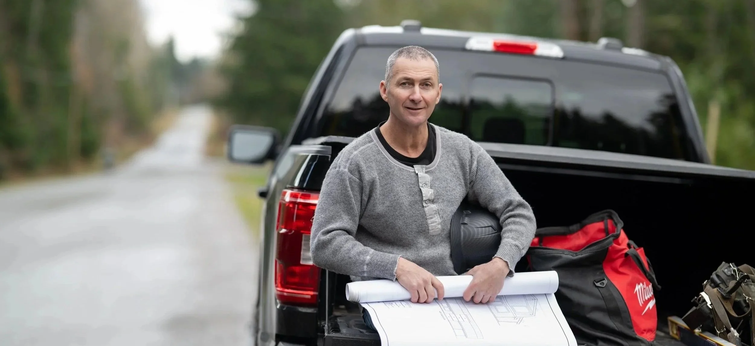 A man standing next to a pickup truck with the tailgate down, holding a rolled-up blueprint, with a red and black tool bag, a black helmet, and other equipment in the truck bed, on a wooded road.