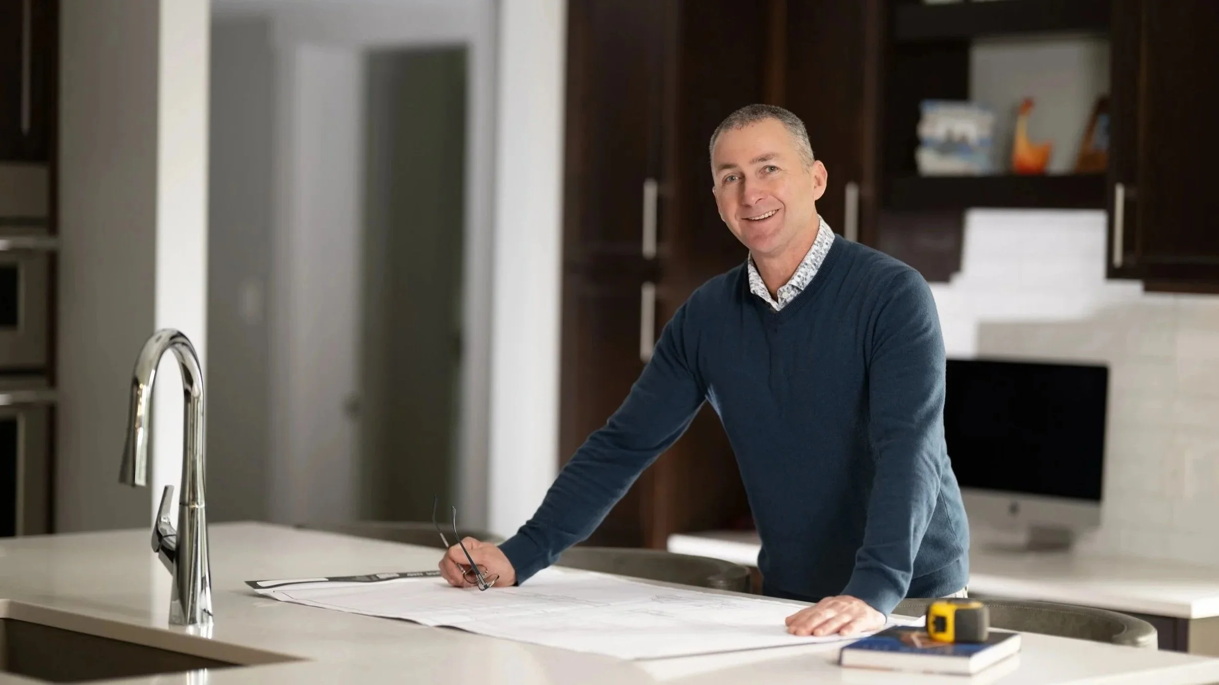 A man stands at a kitchen island, leaning on a large set of blueprints, holding glasses in his right hand, smiling at the camera. He is wearing a blue sweater over a collared shirt. There is a computer monitor, a tape measure, and a book on the island, with a modern kitchen in the background.