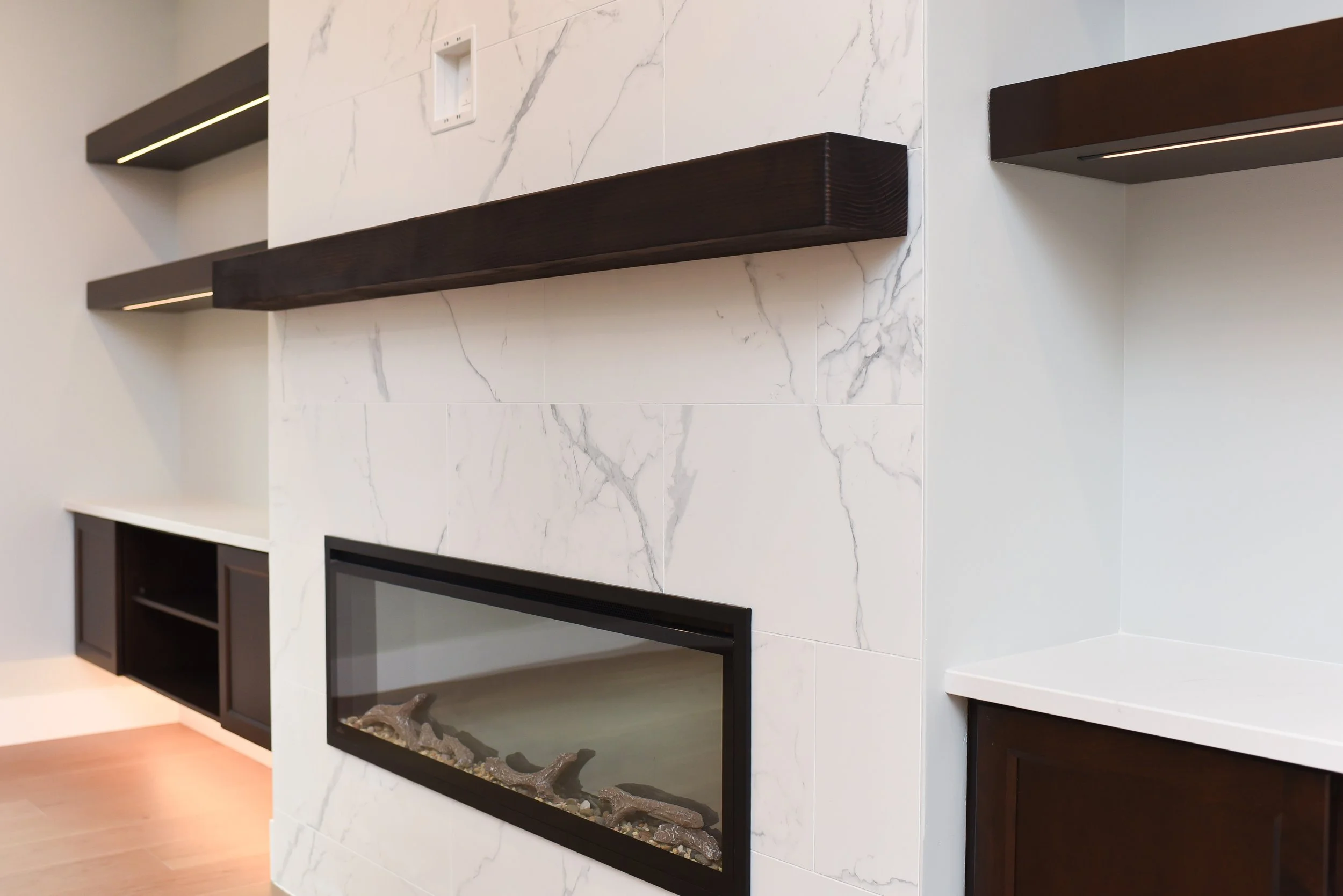Modern interior with a white marble wall with gray veining, dark wood shelves, a built-in fireplace with a black frame and decorative rocks, and a white countertop with dark wood cabinetry.