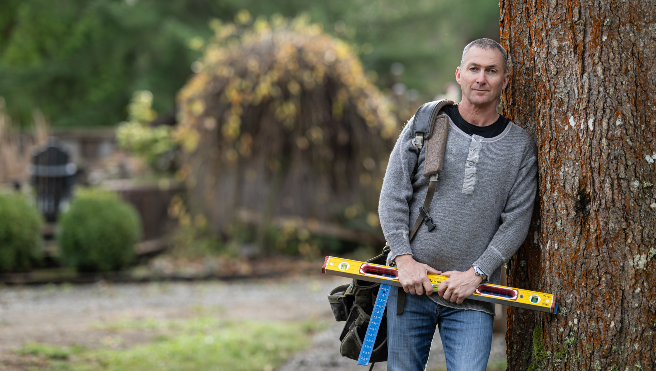 A man leaning against a tree holding a spirit level, outdoors in a park or backyard.