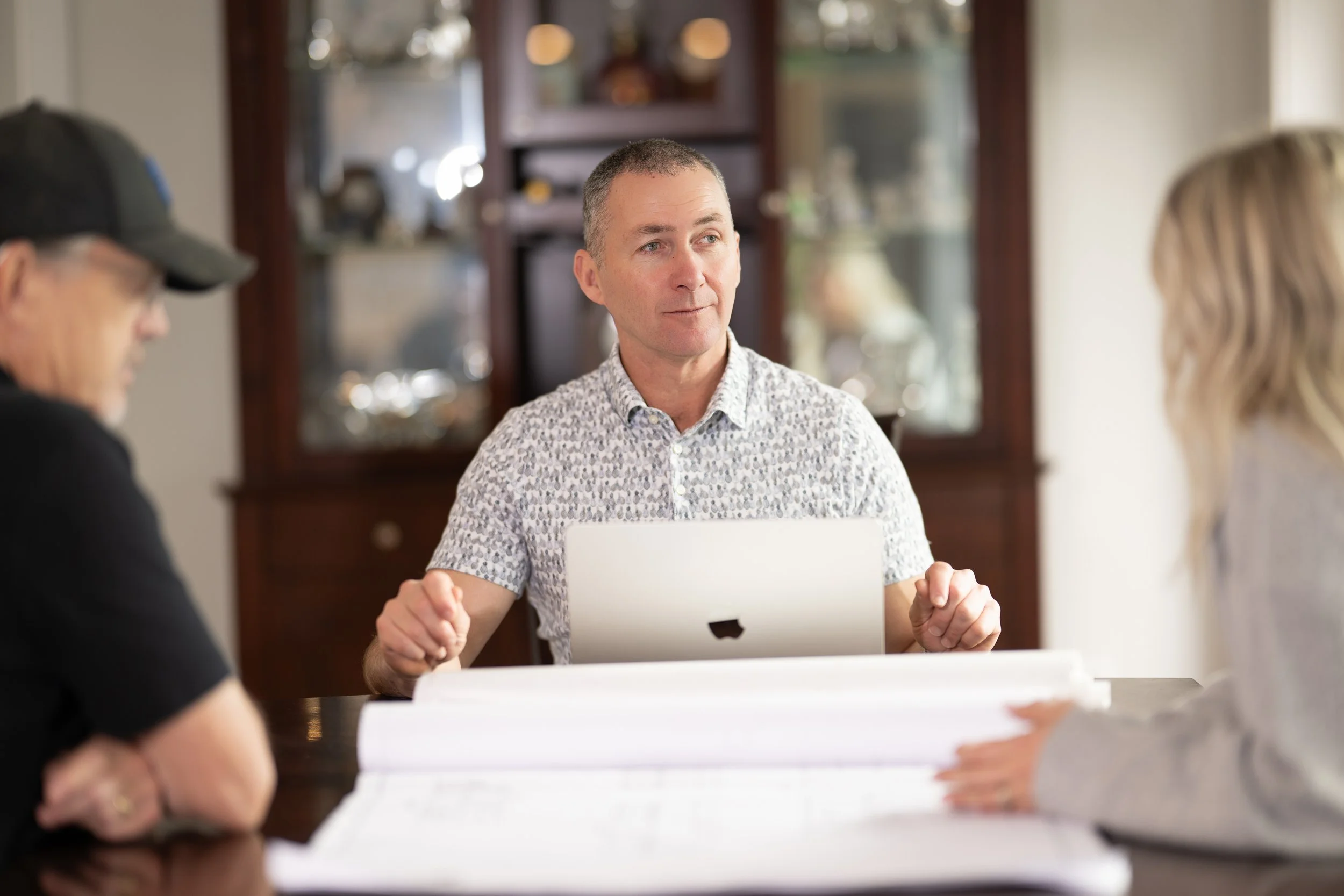 A man sitting at a table with a laptop, talking with two women, one on each side, in a room with wooden furniture and a glass display cabinet behind him.