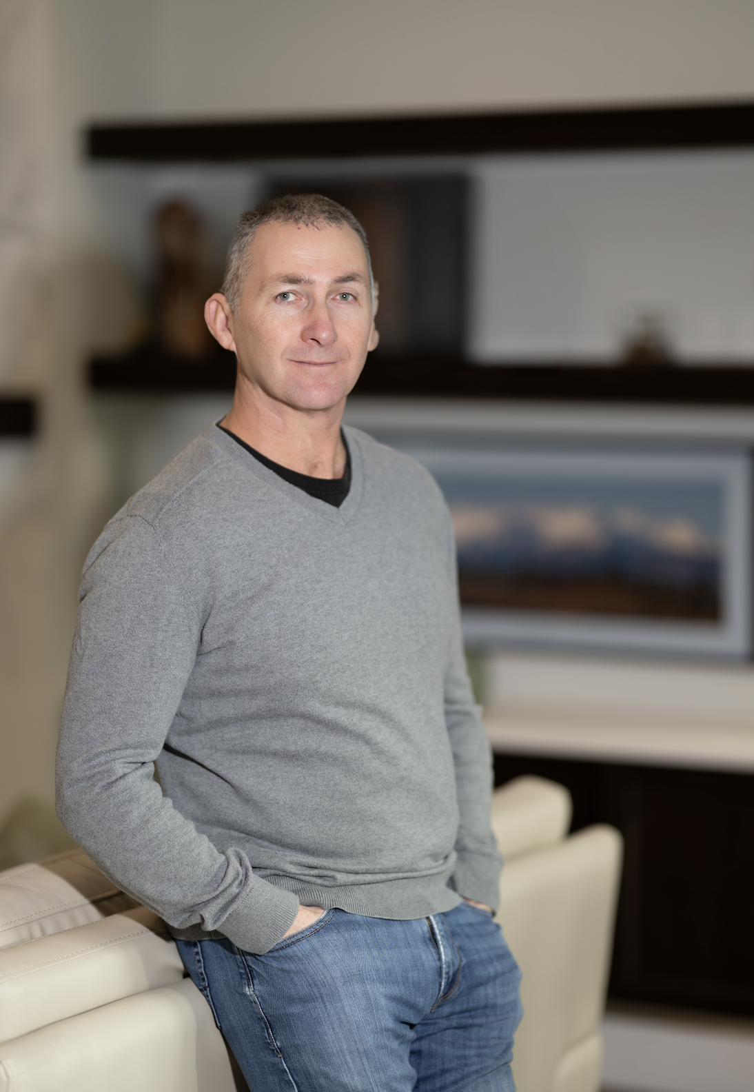 A middle-aged man with short gray hair wearing a gray sweater and jeans, standing indoors with hands in pockets, in front of a beige leather sofa and a blurred background with shelves and a landscape photograph.