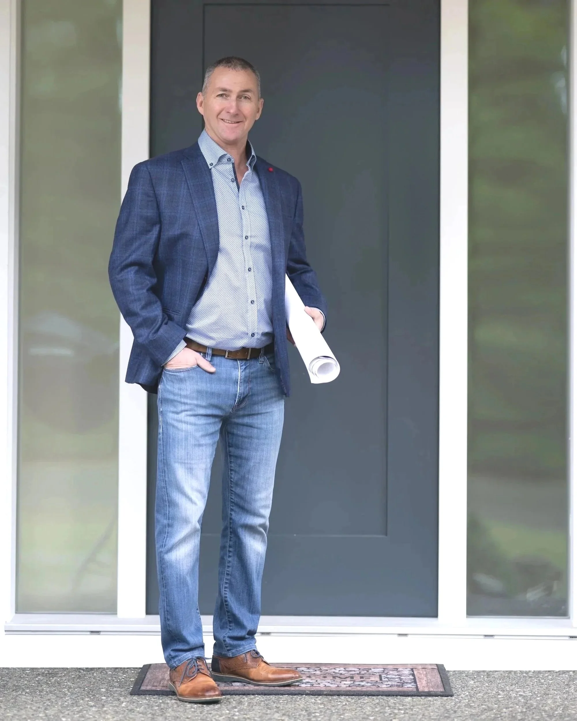A man stands on a front porch holding rolled-up papers, dressed in a blue blazer, light blue button-up shirt, jeans, and brown shoes.