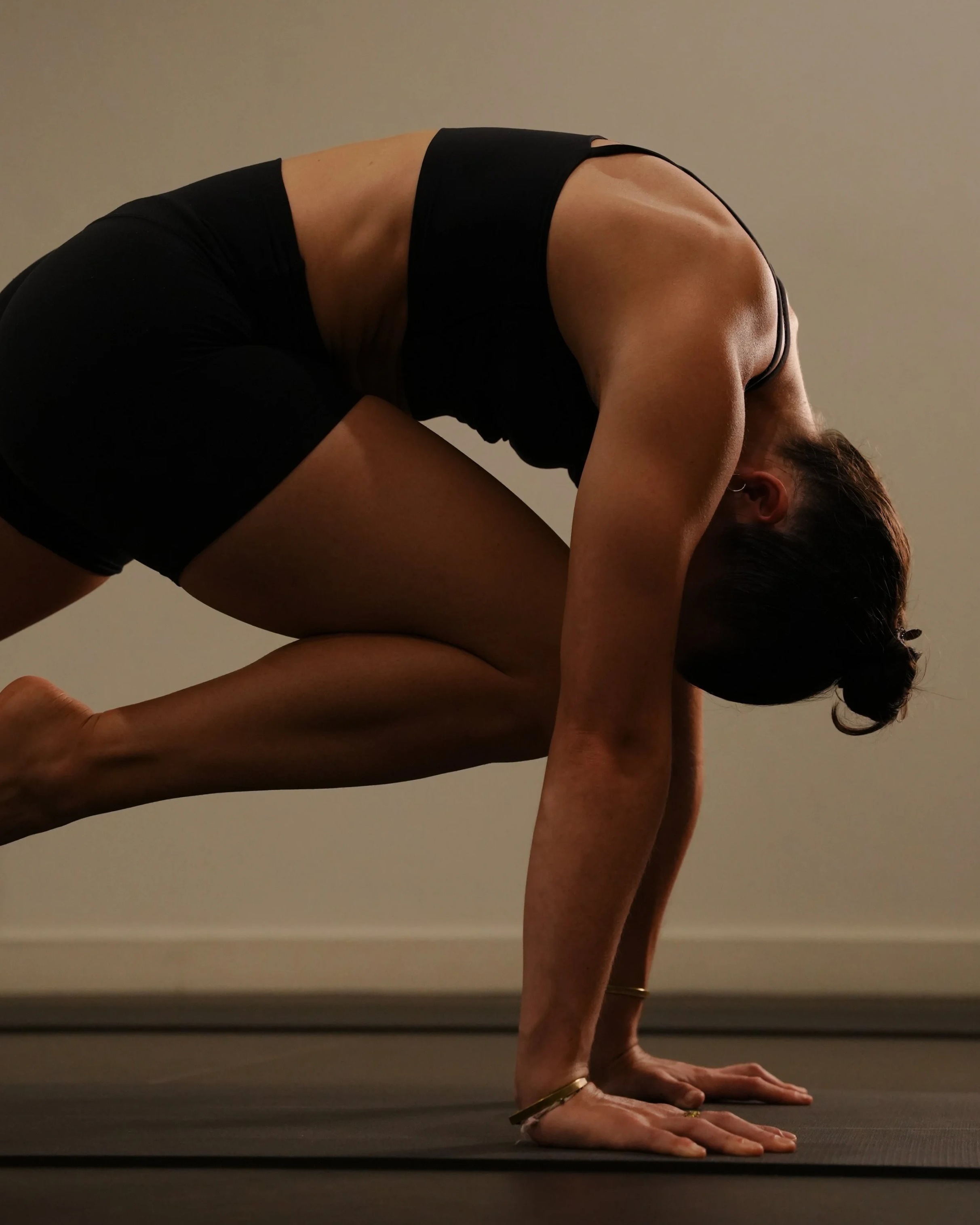 A woman practicing yoga indoors in a crouched forward bend position on a black yoga mat.