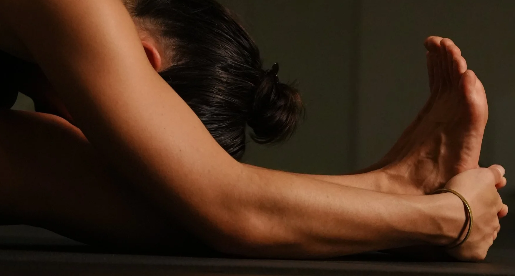 A woman practicing yoga, in a forward bend position with her head resting on her knees and hands together on her feet, in a dimly lit room.