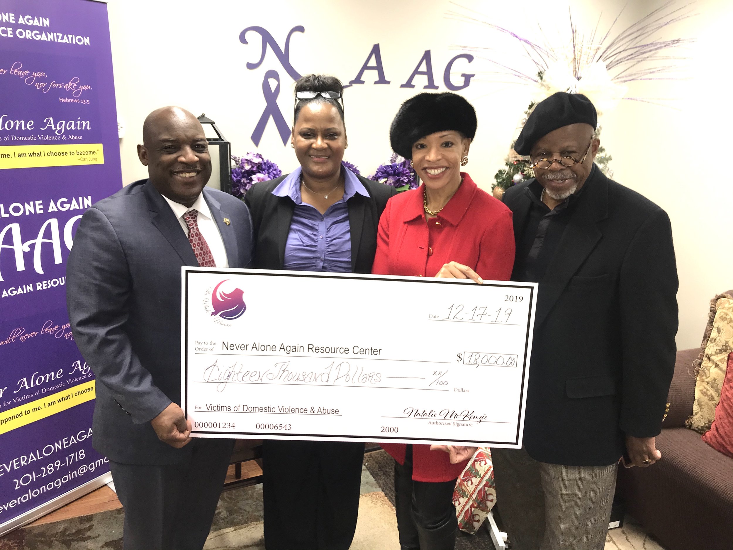 Four people holding a large check for $18,000 made out to Never Alone Again Resource Center, with purple and white organizational banners and purple flowers in the background.