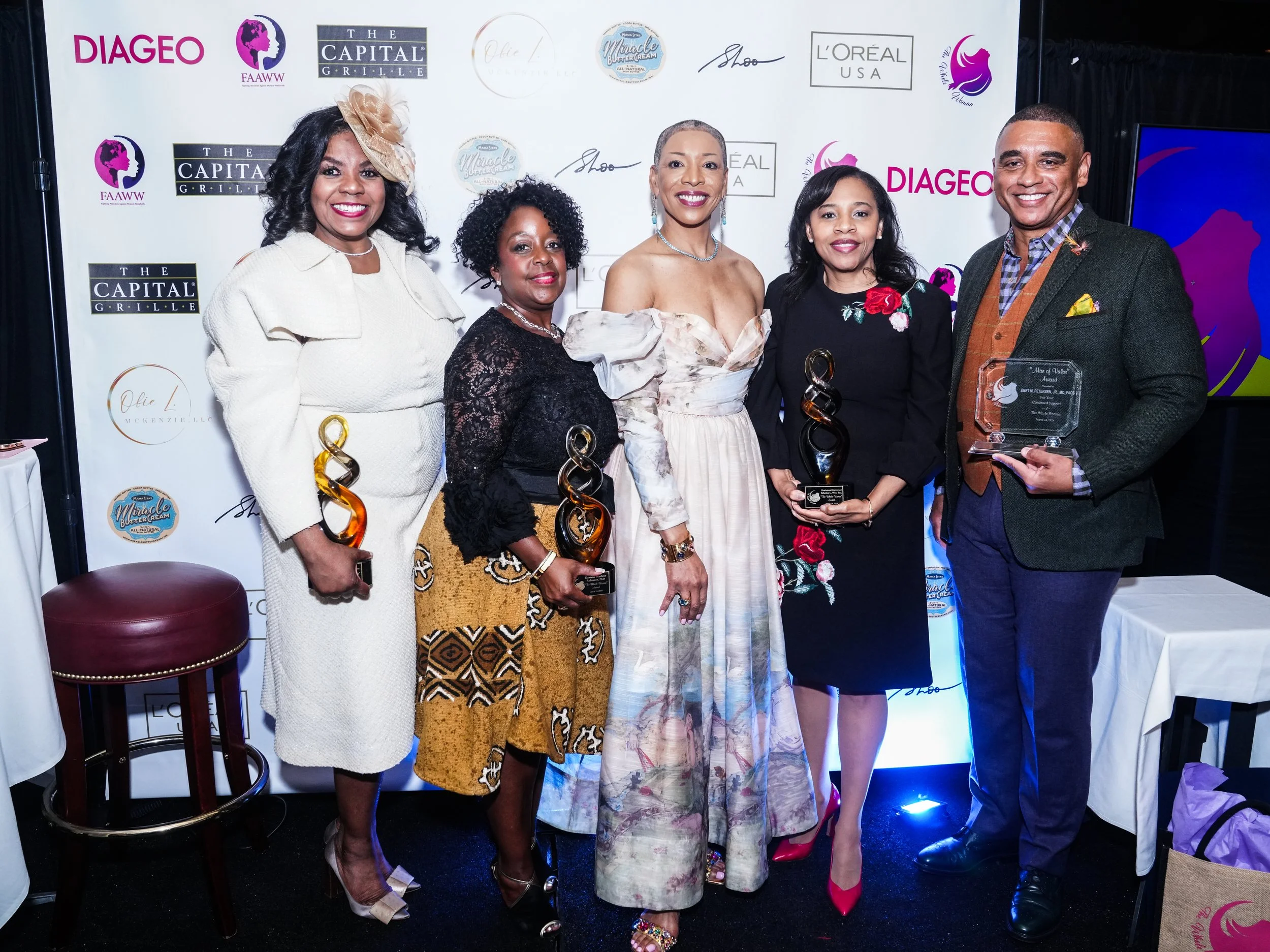 Group of five people at an award ceremony, holding trophies, standing in front of a backdrop with logos, dressed in formal attire.