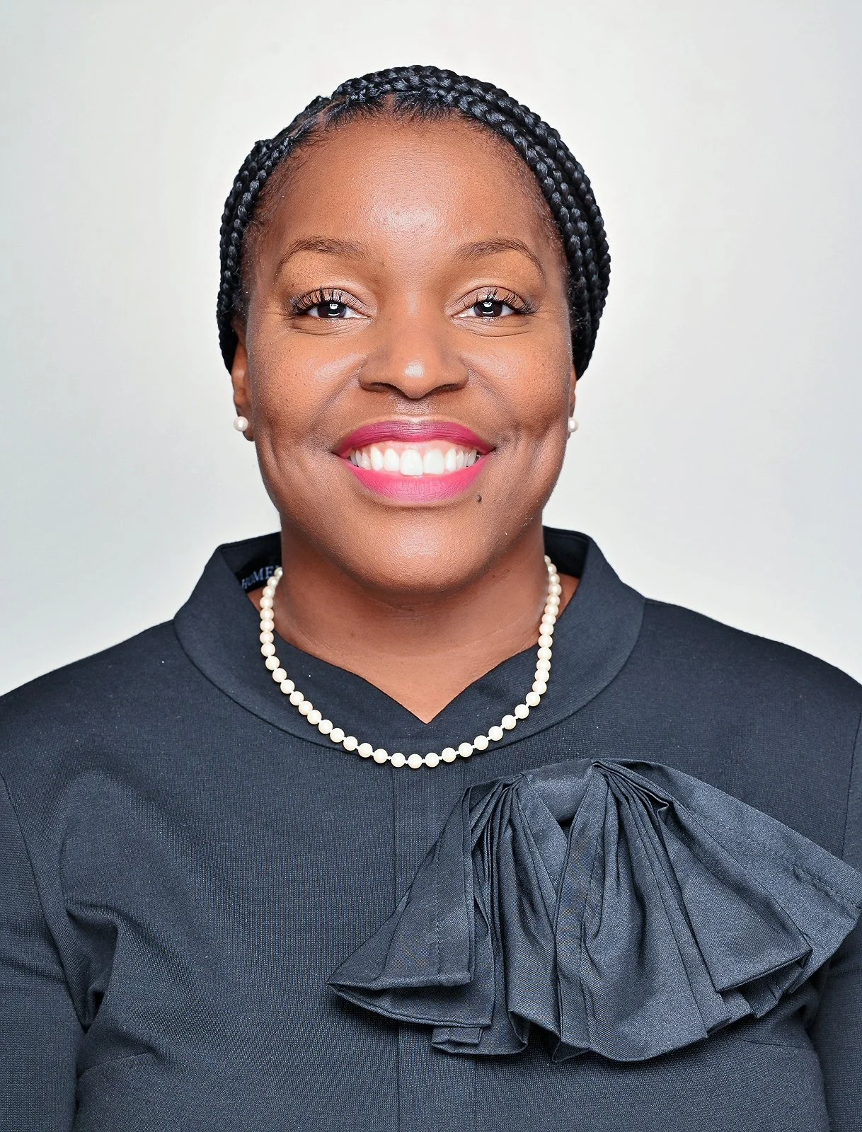 A smiling woman with braided hair, wearing pearl earrings, a pearl necklace, and a black top with a large bow detail.