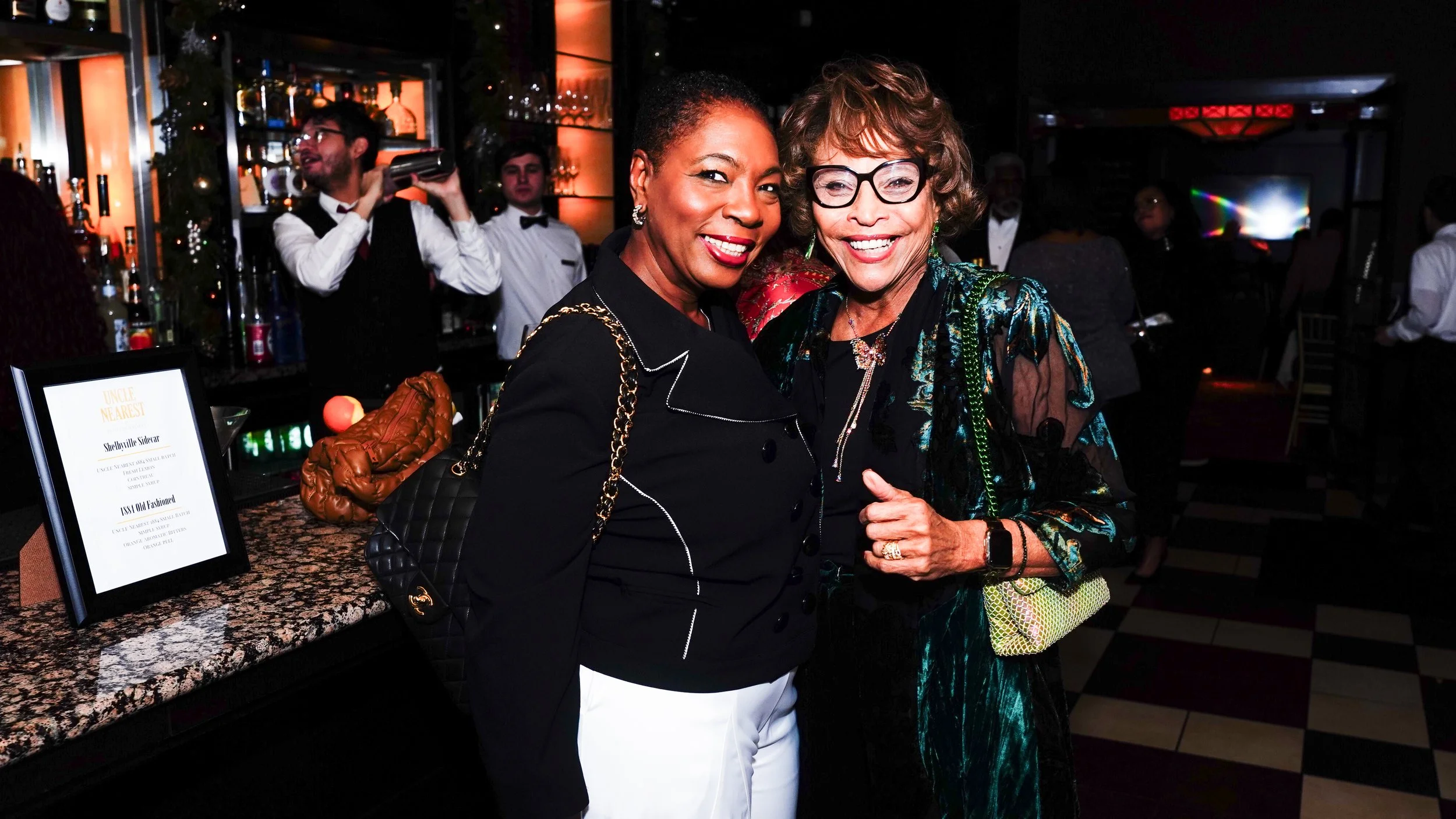 Two women smiling at a social event, standing close together with other guests in the background, in a dimly lit venue with bar and decorations.