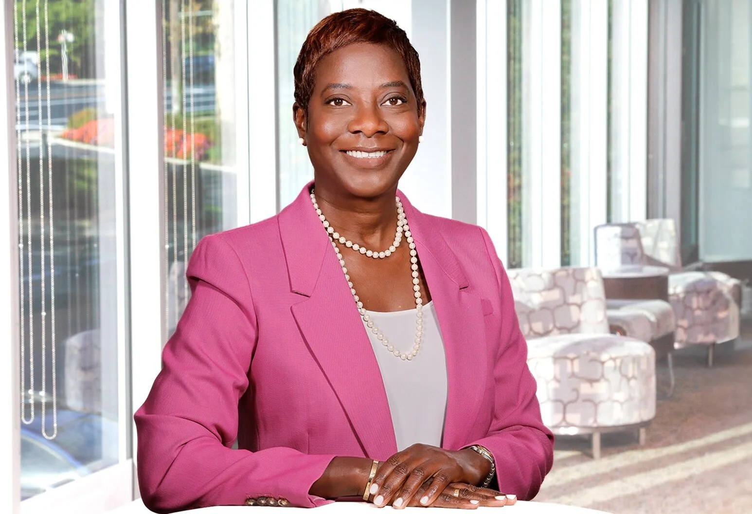 A woman in a pink blazer sitting at a table in a modern office setting with large floor-to-ceiling windows and patterned chairs in the background.