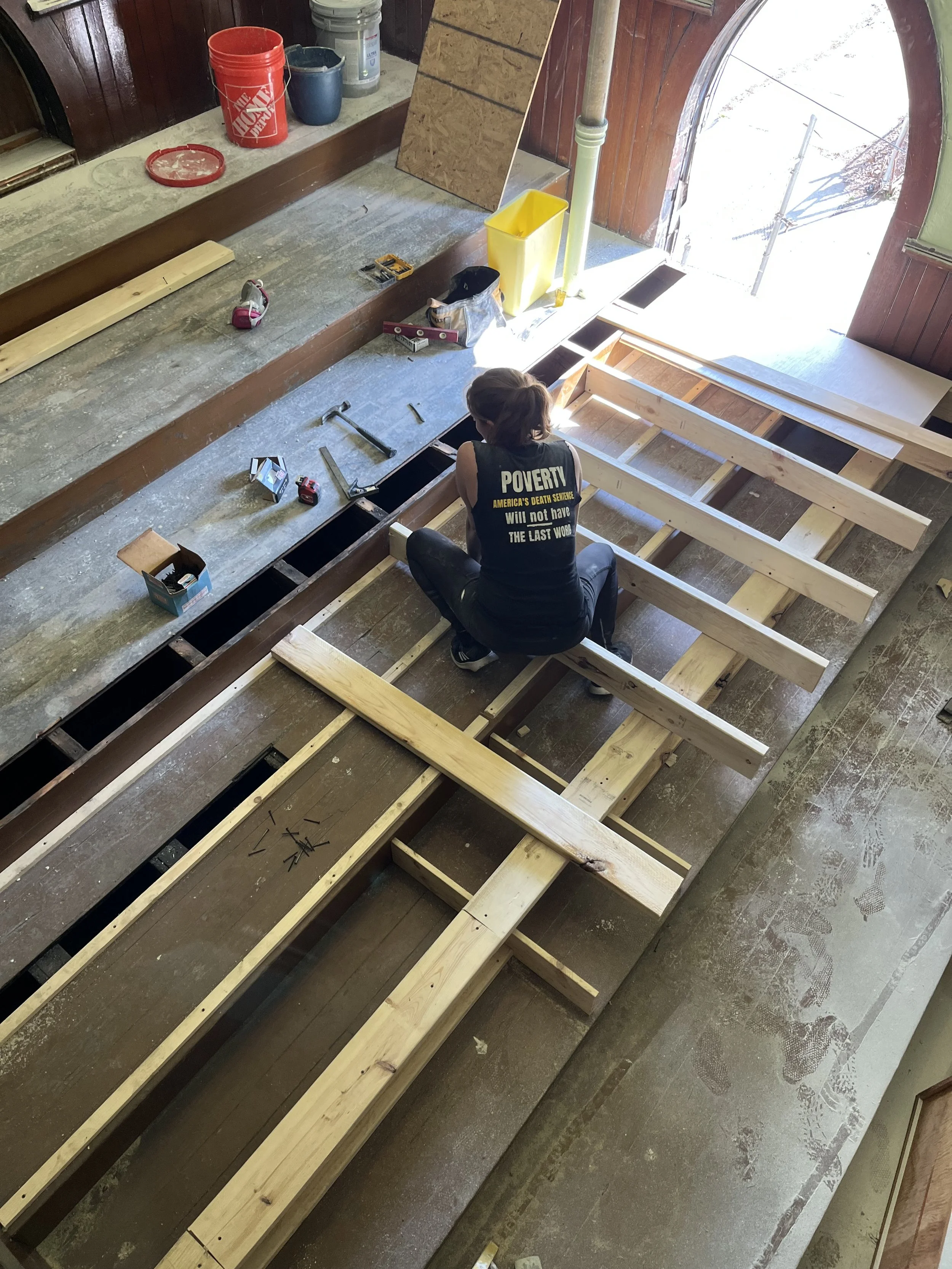 A person building a wooden structure, possibly a floor or platform, inside a workshop or garage. They are sitting on the unfinished frame with tools and materials around. Sunshine streams through a door or window at the open end.