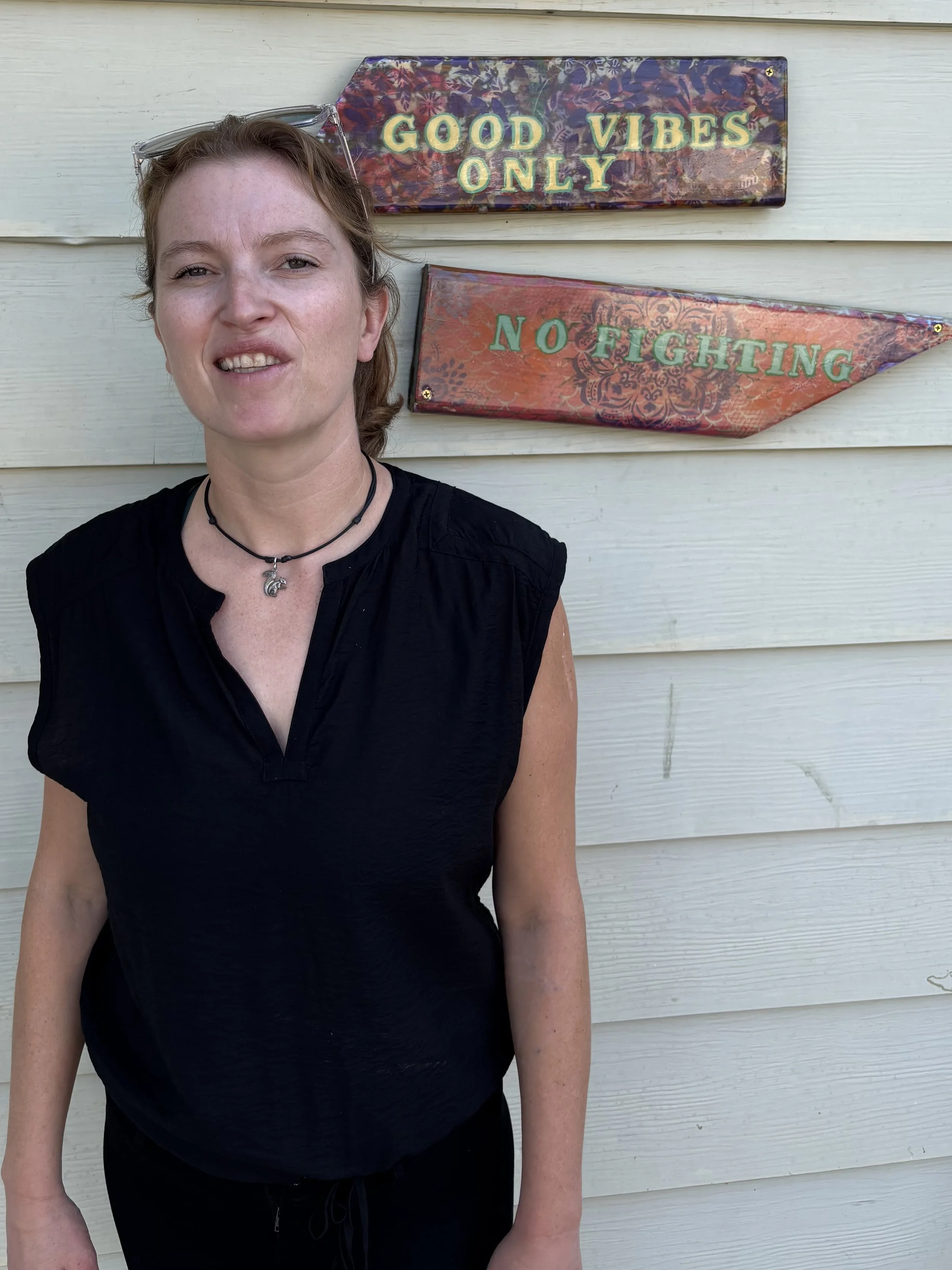 A woman standing in front of a light-colored wooden wall with two decorative signs; one says 'GOOD VIBES ONLY' and the other says 'NO FIGHTING'.