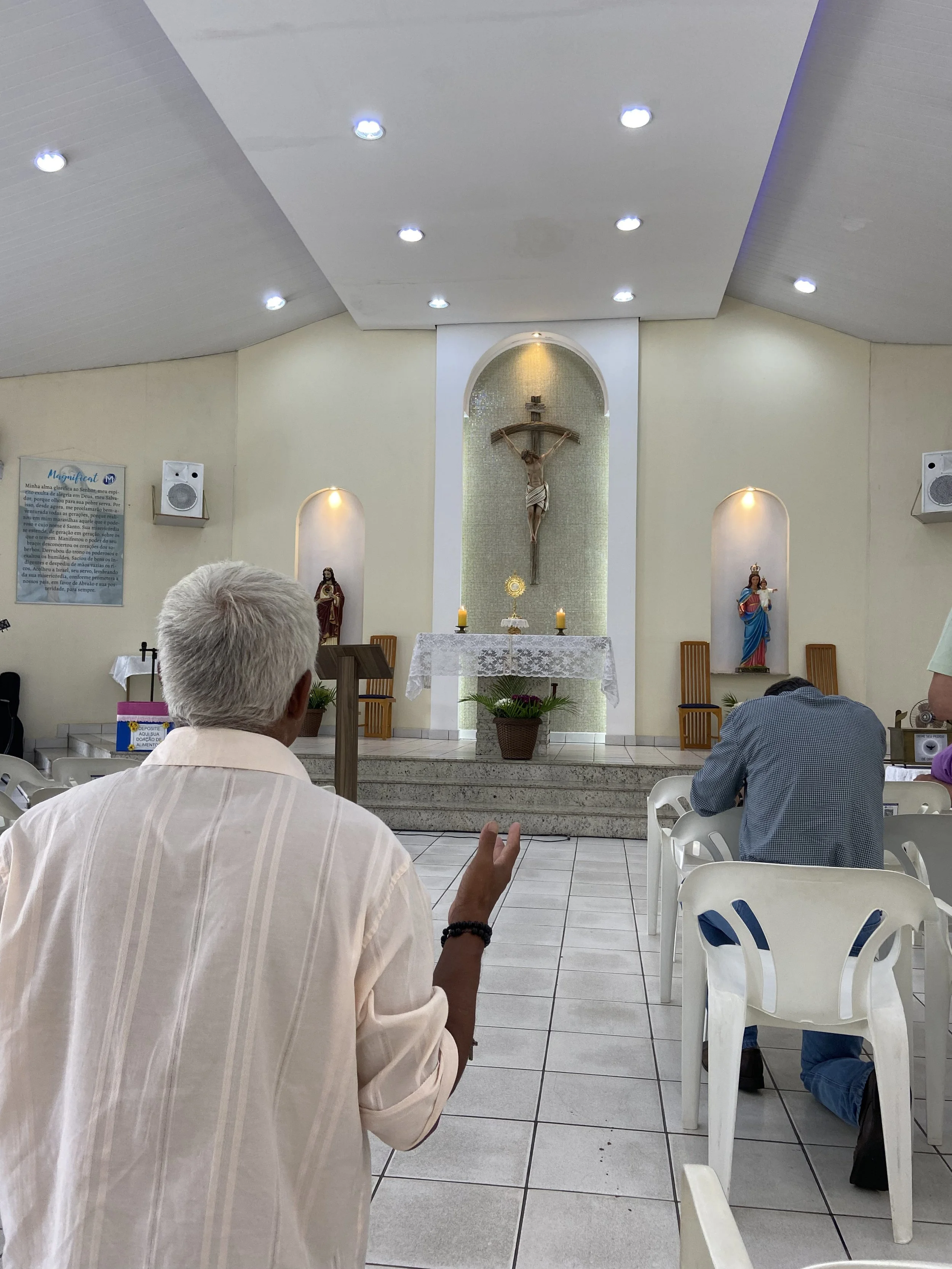 Interior de uma igreja, com pessoas orando ou rezando de costas, ao centro do altar uma imagem de Jesus na cruz, com santuários laterais contendo figuras religiosas, velas acesas e flores.
terço dos homens em sao jose dos campos