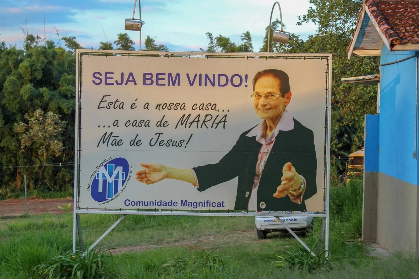 Placa de boas-vindas com uma mulher sorridente, usando jaqueta preta e camisa rosa, gesticulando com as mãos. Texto em português que diz: 'Seja bem vindo! Esta é a nossa casa... a casa de Maria, Mãe de Jesus!'. Logo na parte inferior esquerdo, o nome da comunidade: 'Comunidade Magnificat'.