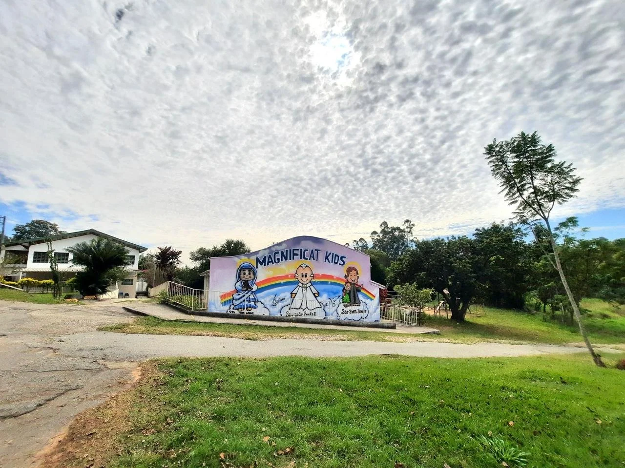 Painel de uma escola infantil chamada Magnificat Kids com desenhos de personagens infantis, arco-íris e árvores ao redor.