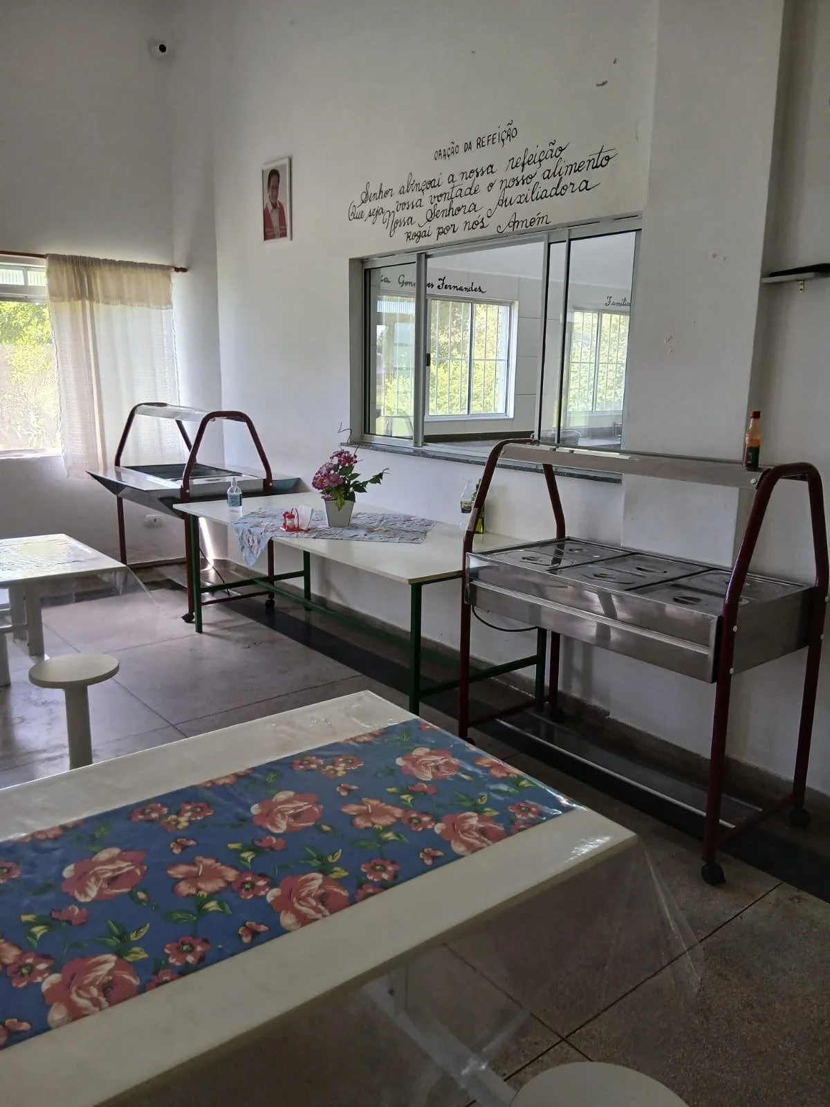 Sala de refeitório com mesa coberta por toalha com estampa de flores, uma planta com flores artificiais na mesa ao centro, duas carrinhos de comida vazios, janela ao fundo com grades e cortina, parede branca com frase escrita em português e retrato d