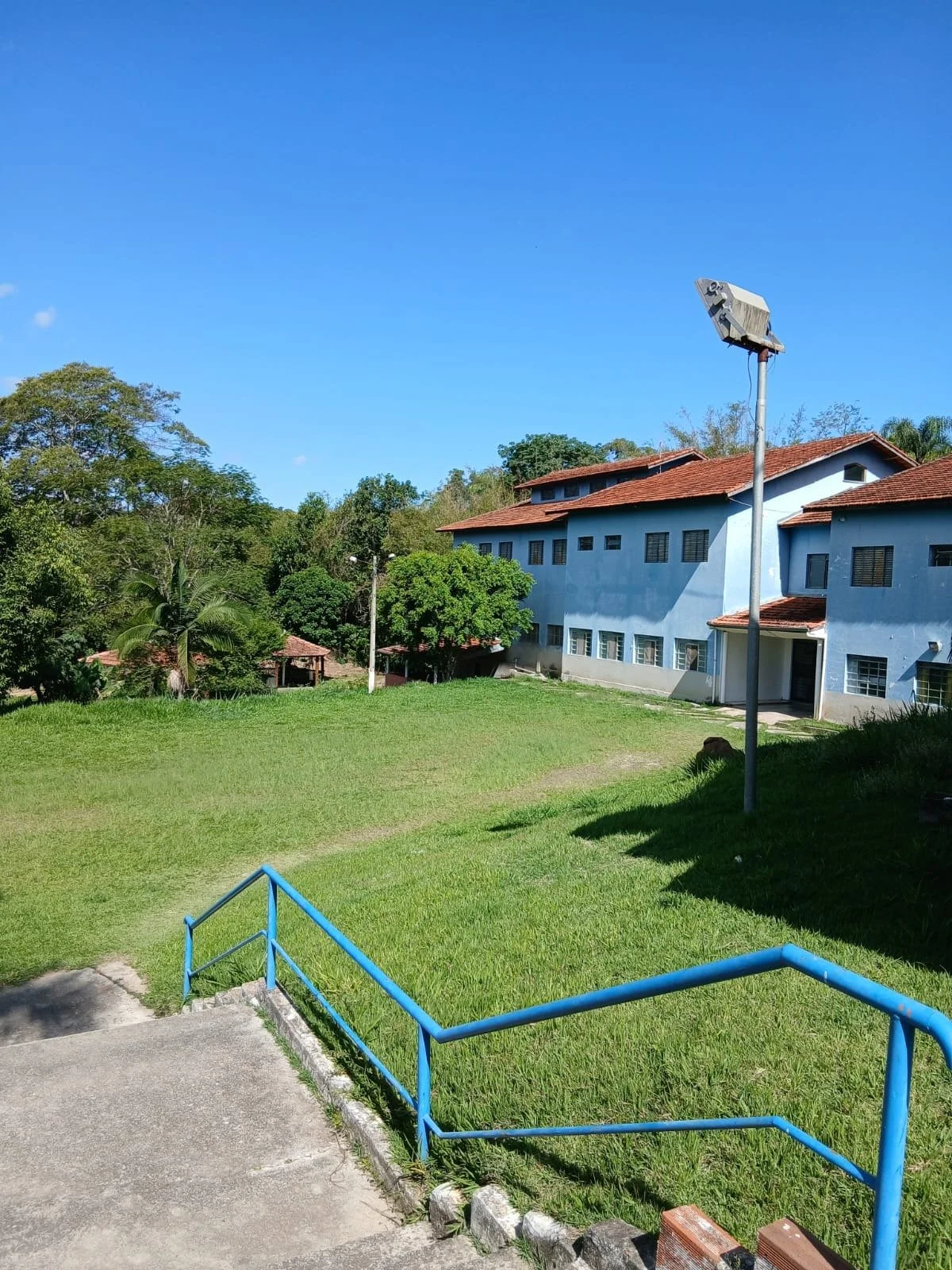 Área verde com gramado, uma escadaria com guarda-corpo azul, uma casa azul com telhado vermelho ao fundo, árvores ao redor e um poste de iluminação.