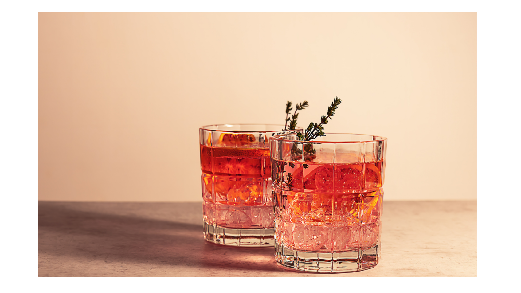 Two glasses of pink cocktail garnished with sprigs of thyme on a wooden surface against a neutral background.