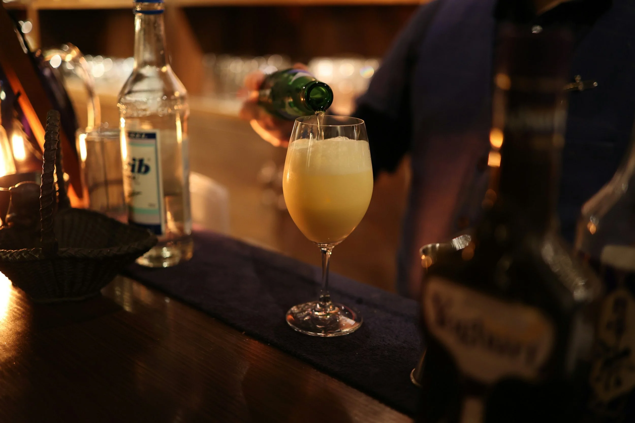 A bartender pouring a drink into a champagne flute on a dark bar counter, with bottles and bar tools in the background.