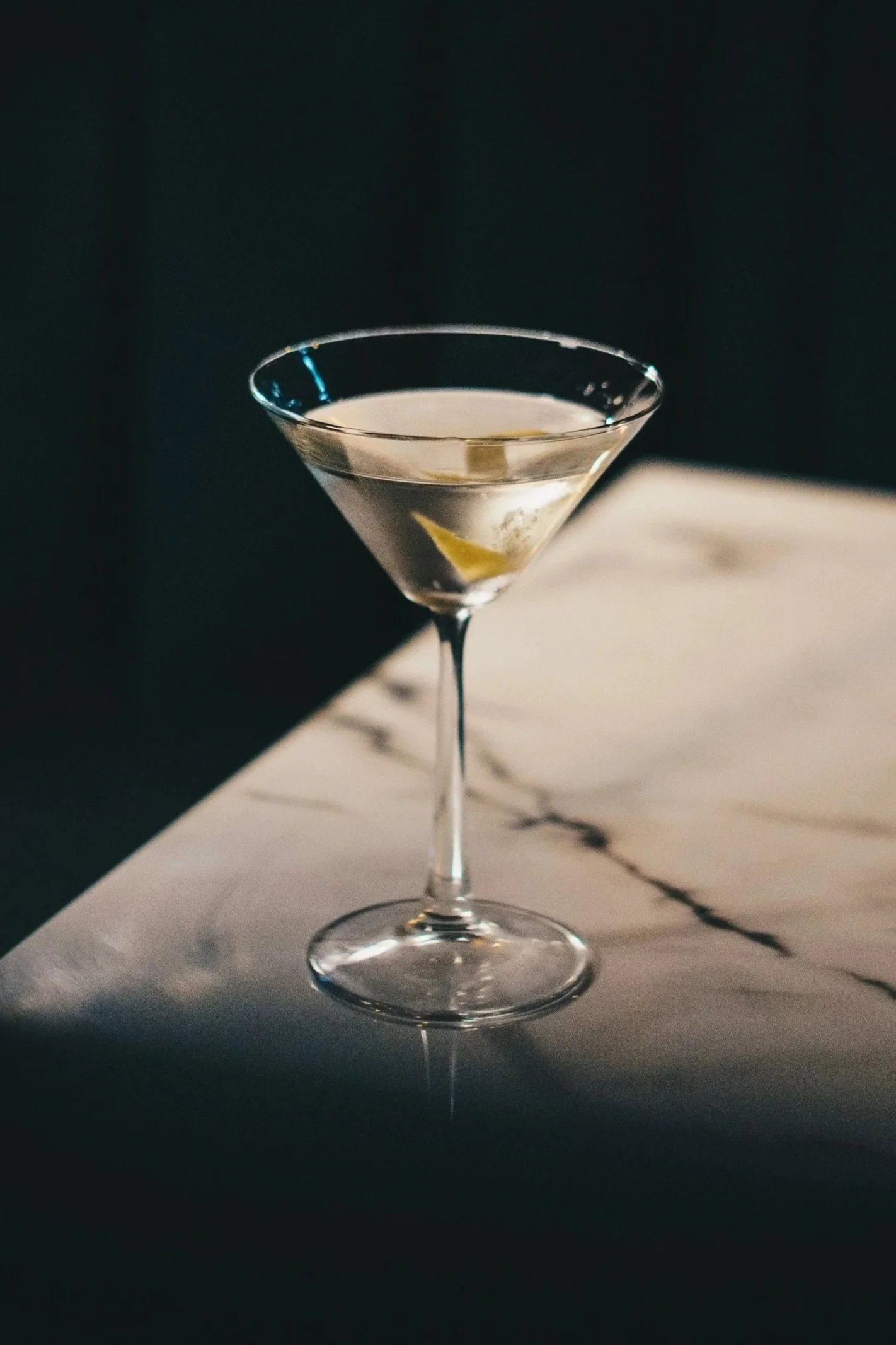 A martini glass filled with a clear cocktail garnished with a lemon twist, placed on a marble surface against a dark background.