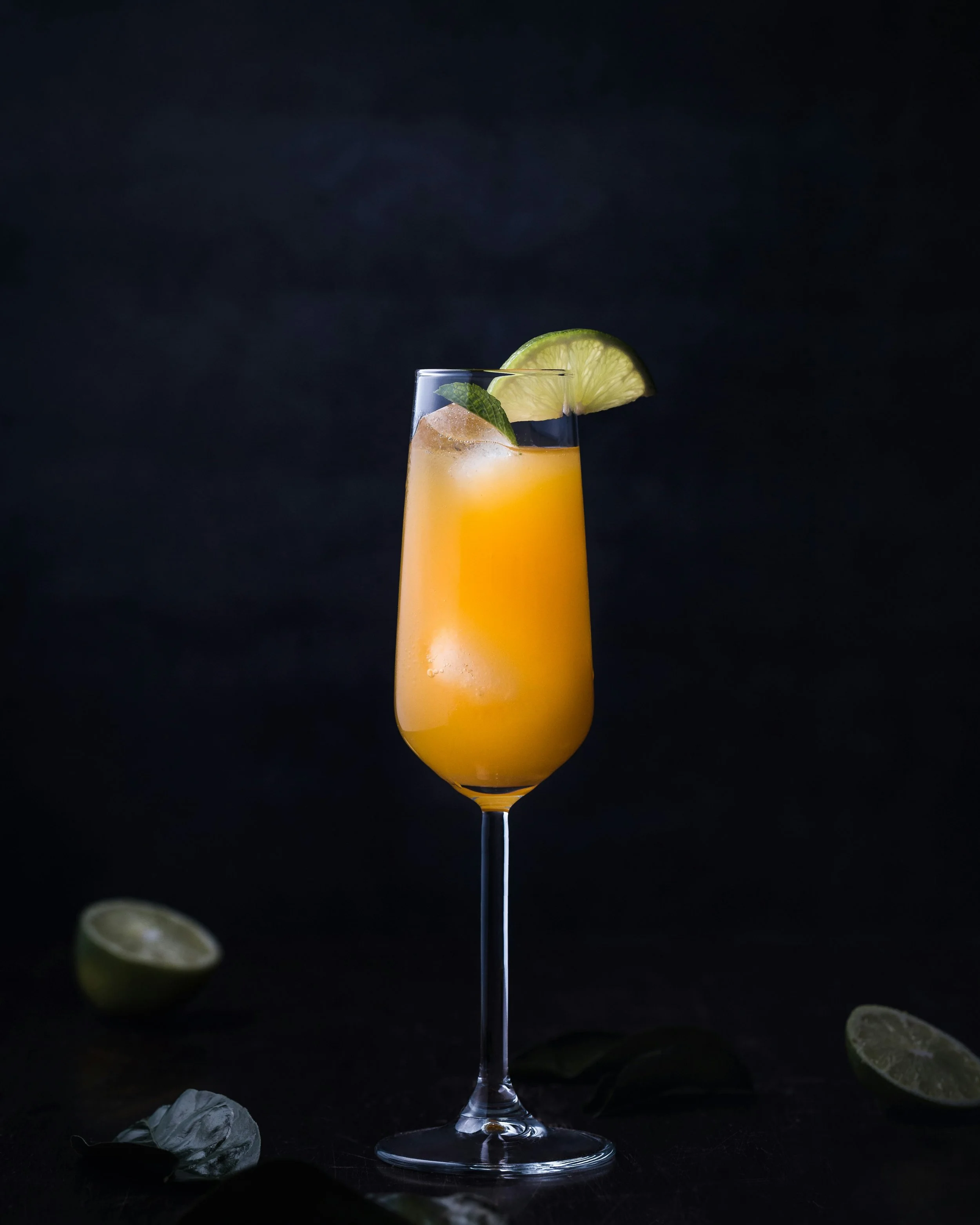 A tall flute glass filled with orange cocktail ice and garnished with lime wedge and mint leaves, on a black background with lime slices and leaves scattered around base.