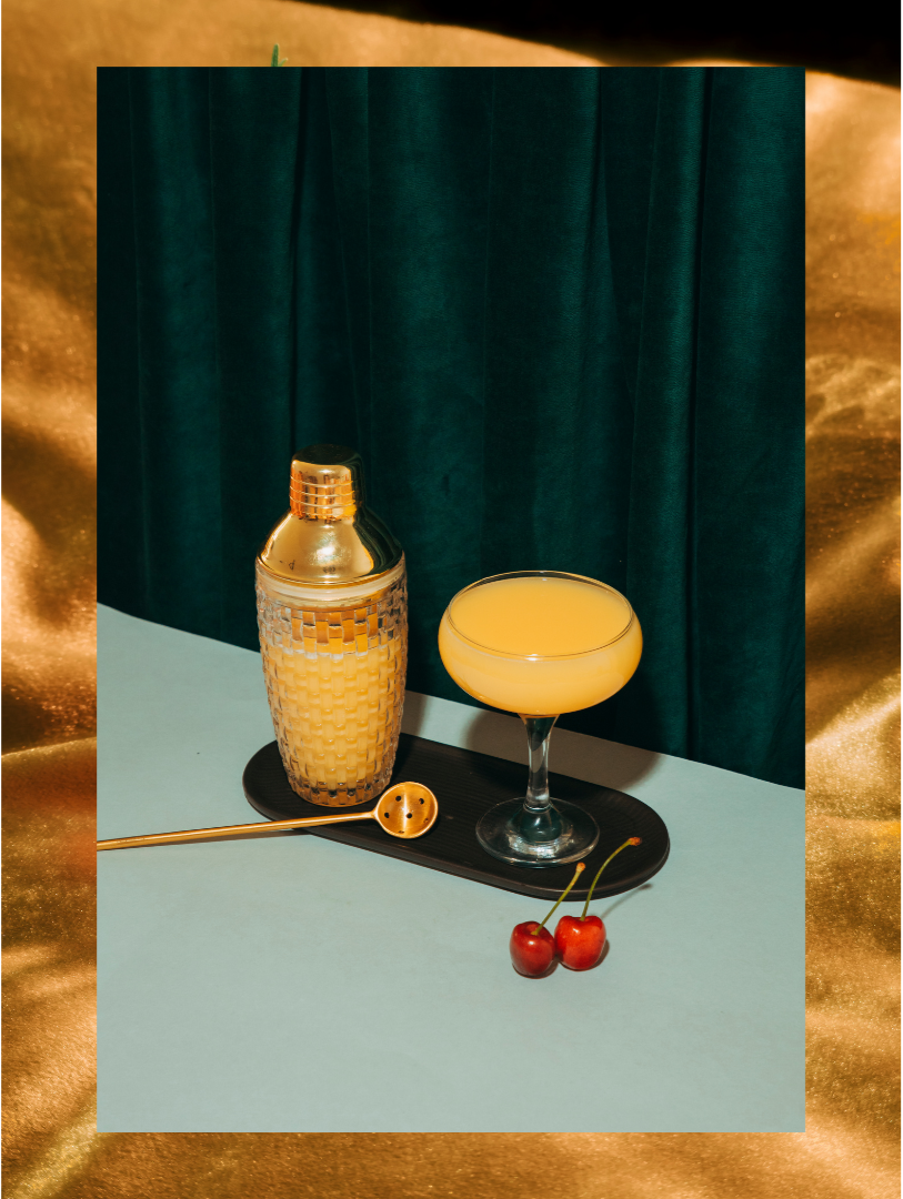 A gold cocktail shaker, a coupe glass filled with a yellow cocktail, and two cherries on a light blue surface with a dark wood tray underneath the glass, set against a teal curtain background.