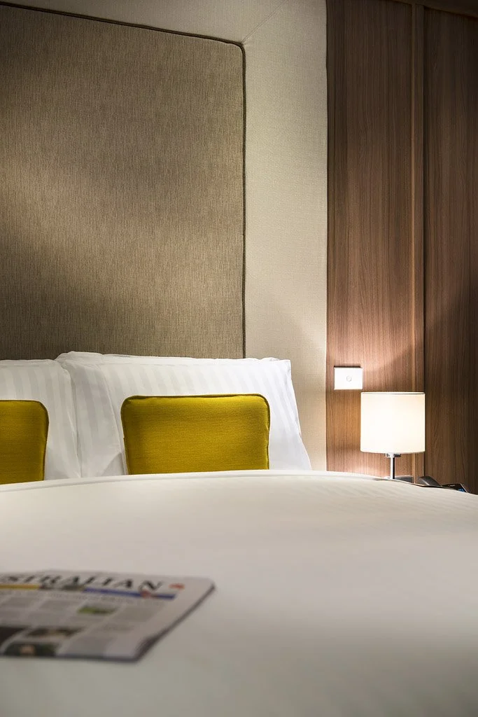 A hotel bed with white linens and two white pillows with yellow decorative pillows behind them, a beige headboard, a wooden wall with a lamp attached, and a newspaper on the bed.