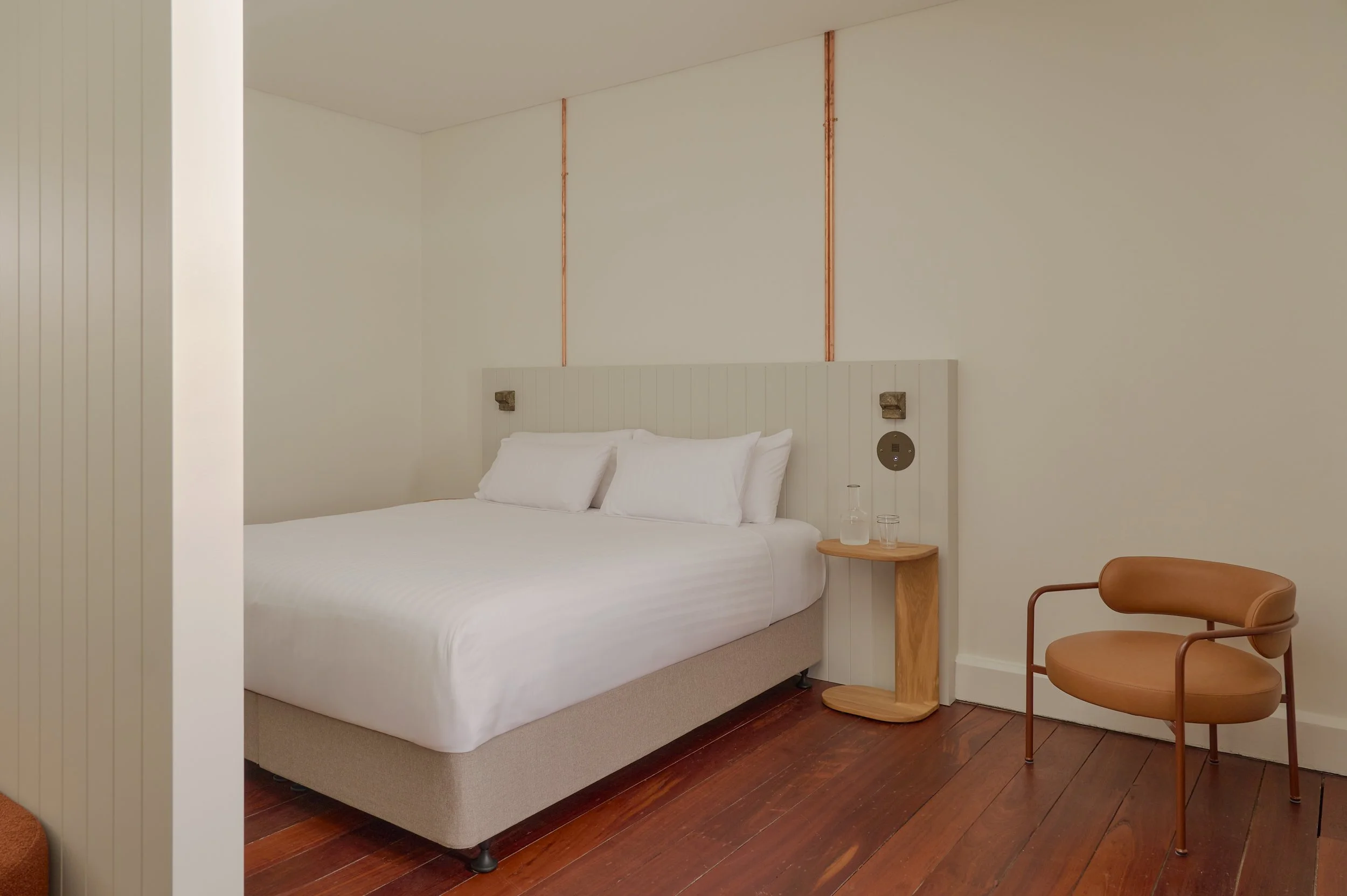 A modern bedroom with a white bed, a wooden side table with glass water bottles, and a tan chair on wooden flooring.
