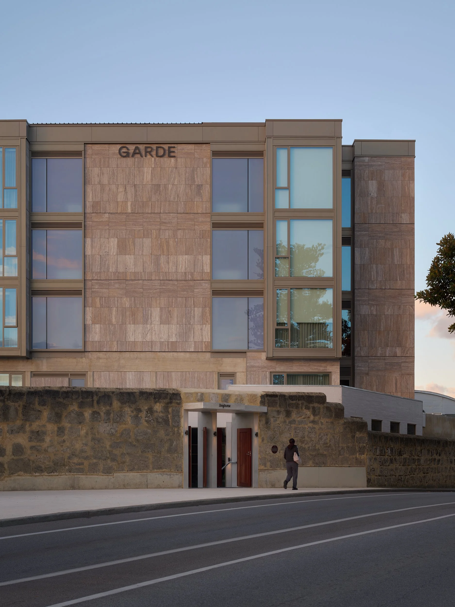 Modern multi-story building with the sign 'GARDE,' large rectangular windows, and a stone wall entrance. A sidewalk and a person walking are in the foreground, with a clear blue sky in the background.