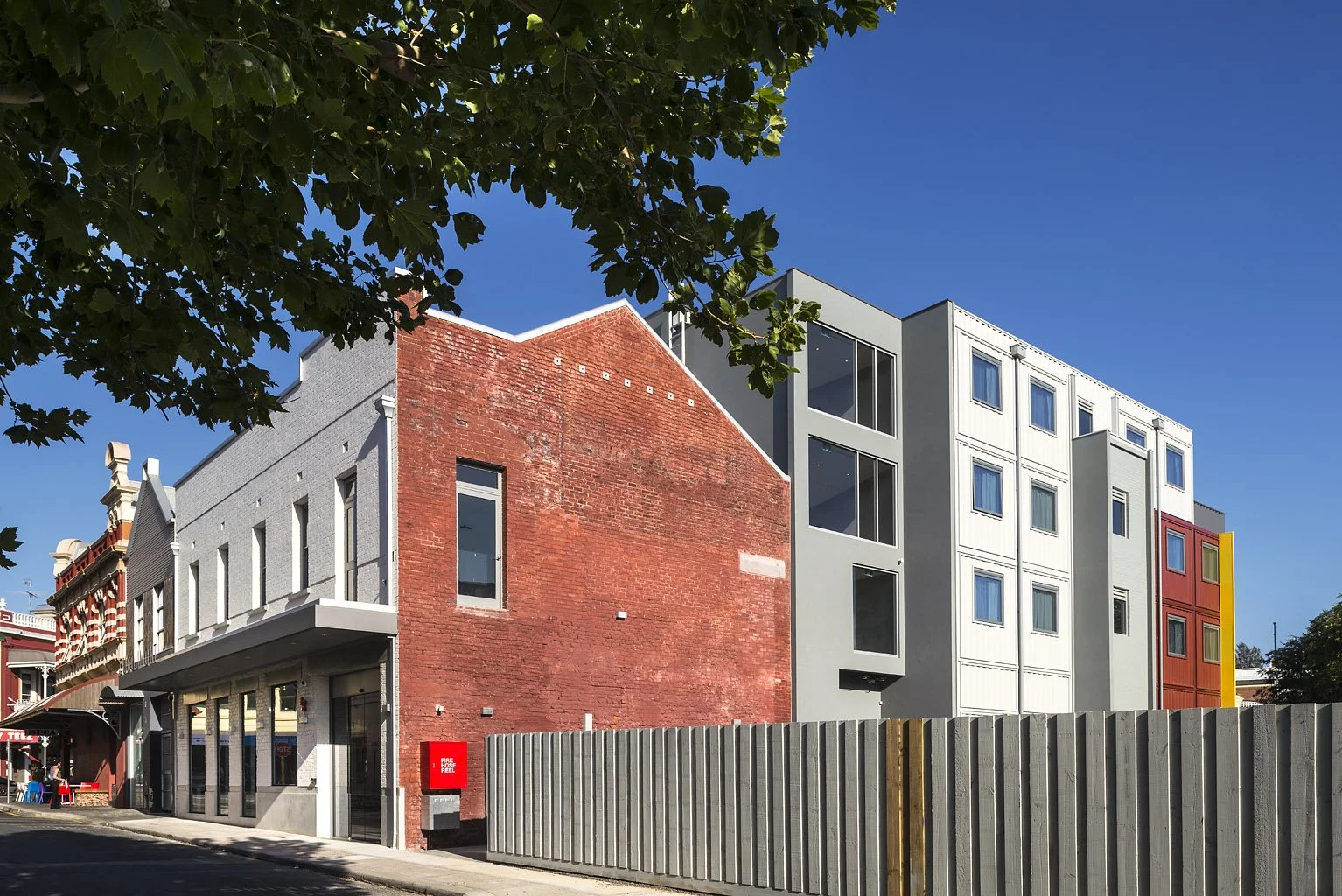 A multi-story building with a mixture of architectural styles. The left side has an older brick facade with arched windows, while the right side features a modern gray and colorful exterior with large windows. A fence runs along the street in front, 