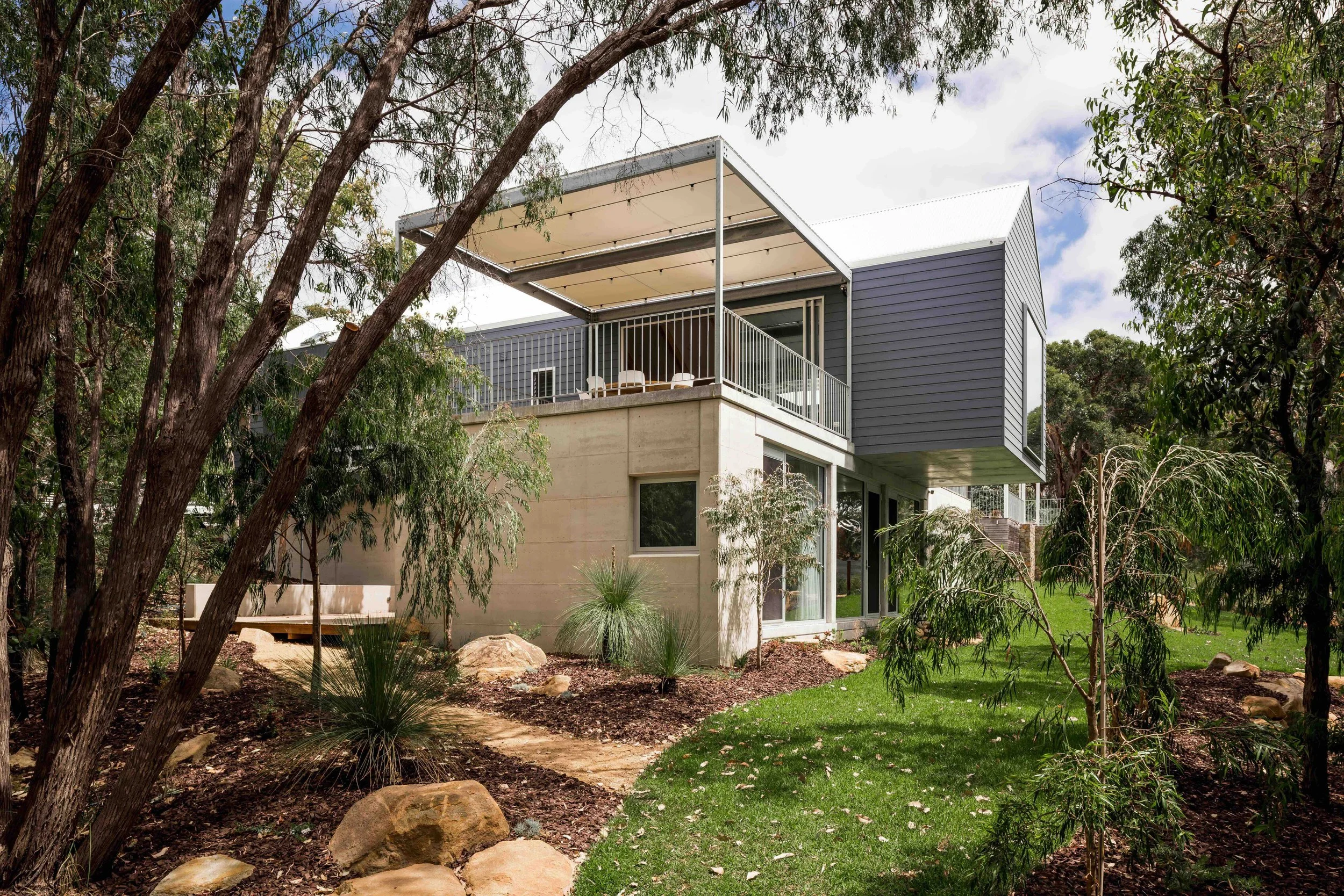 Modern house with a concrete lower level and a gray upper level, surrounded by trees and landscaped yard with rocks and green grass.