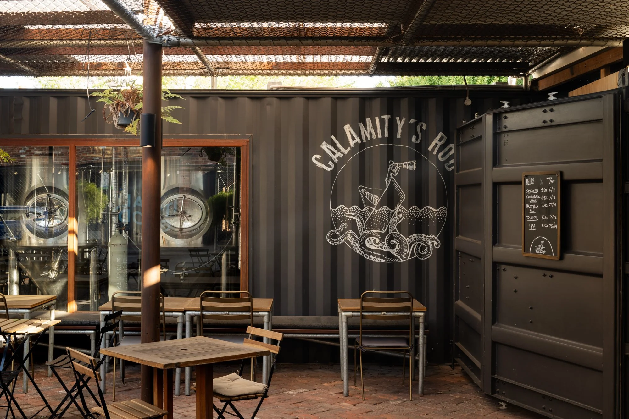 Indoor seating area with wooden tables and chairs, a black wall featuring a logo of a boat and an octopus with the text "Calamity's Ruo" and a menu listing beers.