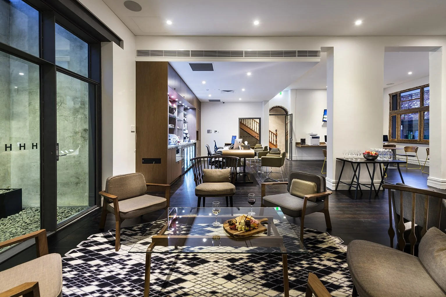 Interior of a modern lounge area with seating, a glass coffee table with food and drinks, and a background area with a buffet and computer stations.