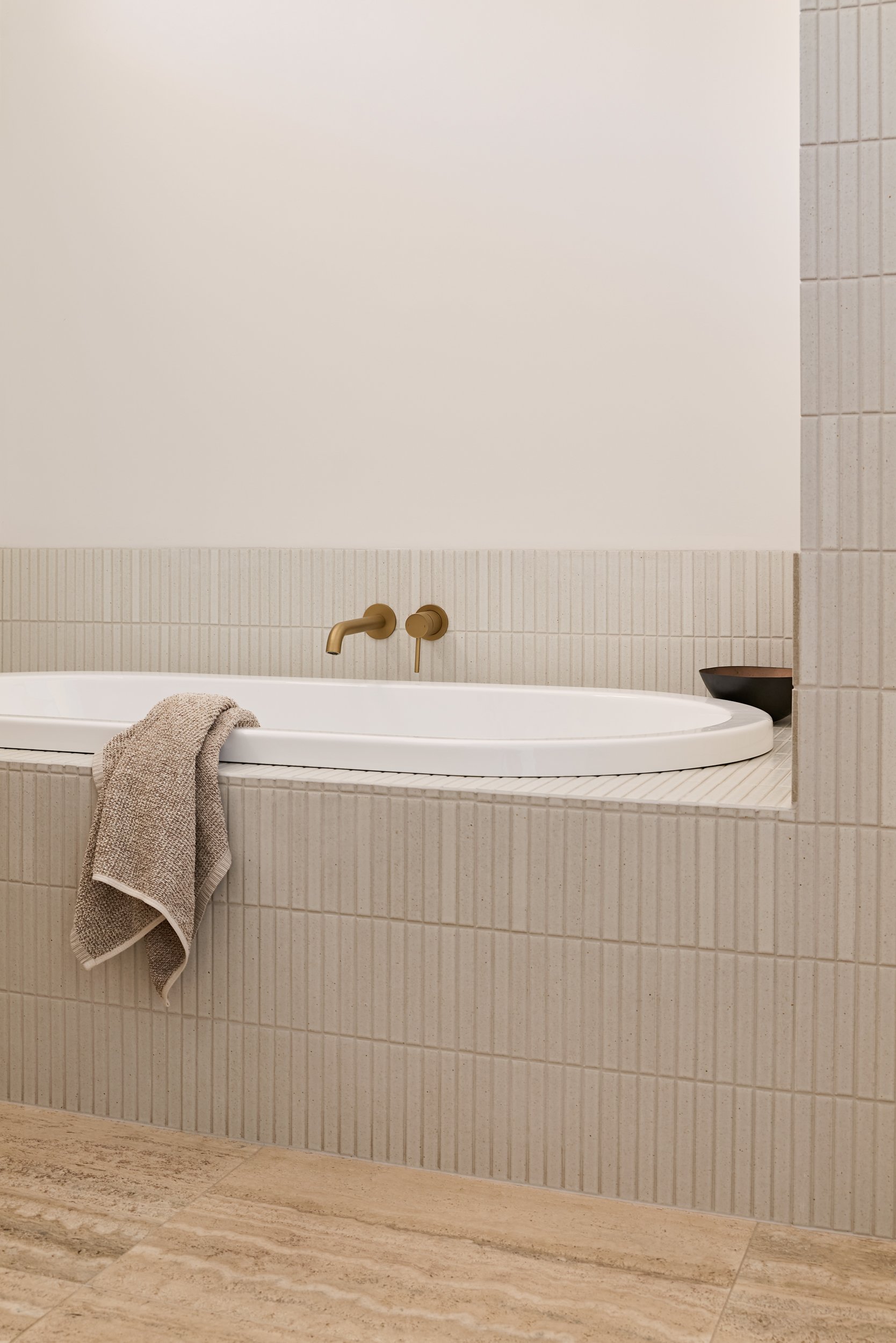 Bathroom with built-in bathtub, brass faucet, wall-mounted controls, beige tiled walls, and a towel hanging on the side.