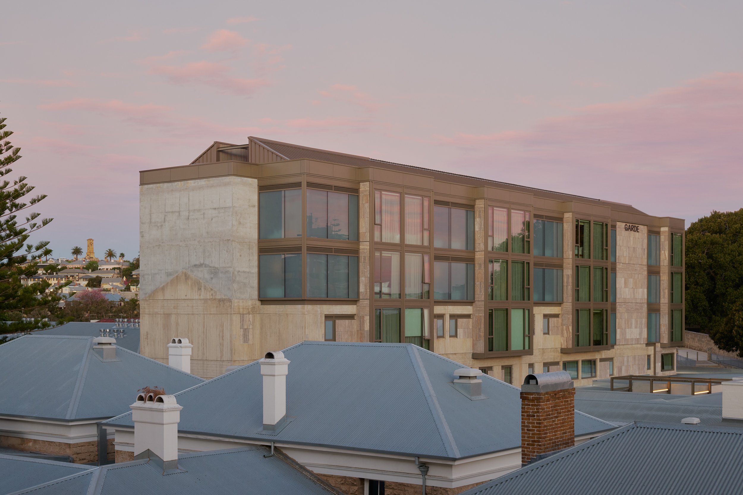 Modern multi-story building with large windows, beige stone facade, and a metal roof in a suburban neighborhood at sunset.