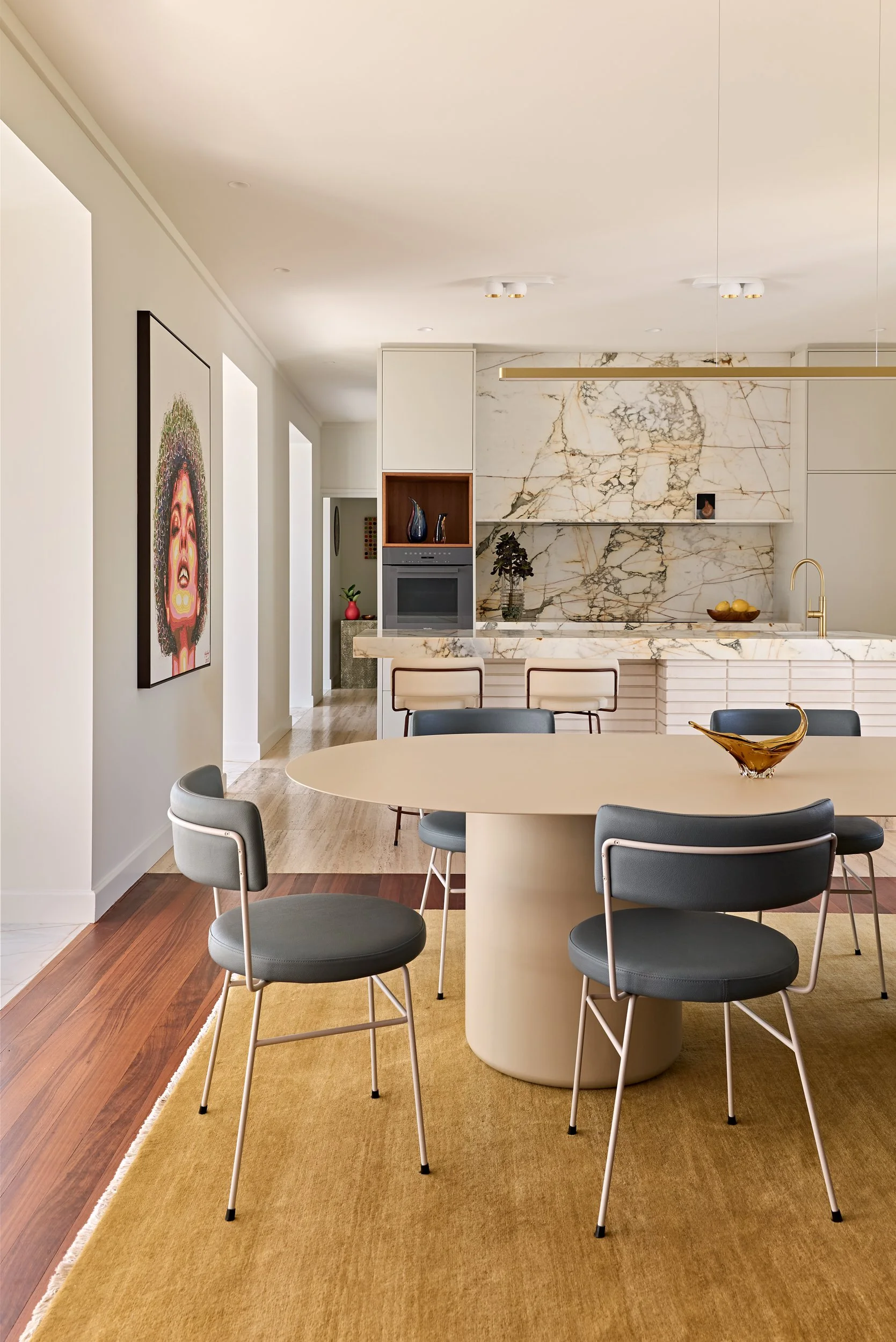 Modern dining area with a round cream-colored table, six gray and black chairs, and a yellow rug. In the background, a kitchen with marble countertops and a backsplash, a built-in oven, and metallic fixtures.