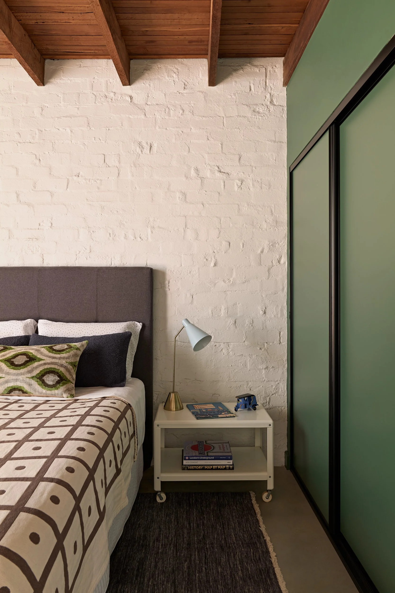 A bedroom with a brown bed with patterned pillows, a white side table with a lamp, books, and a toy truck, a white brick wall, a green closet door, and a wooden ceiling.