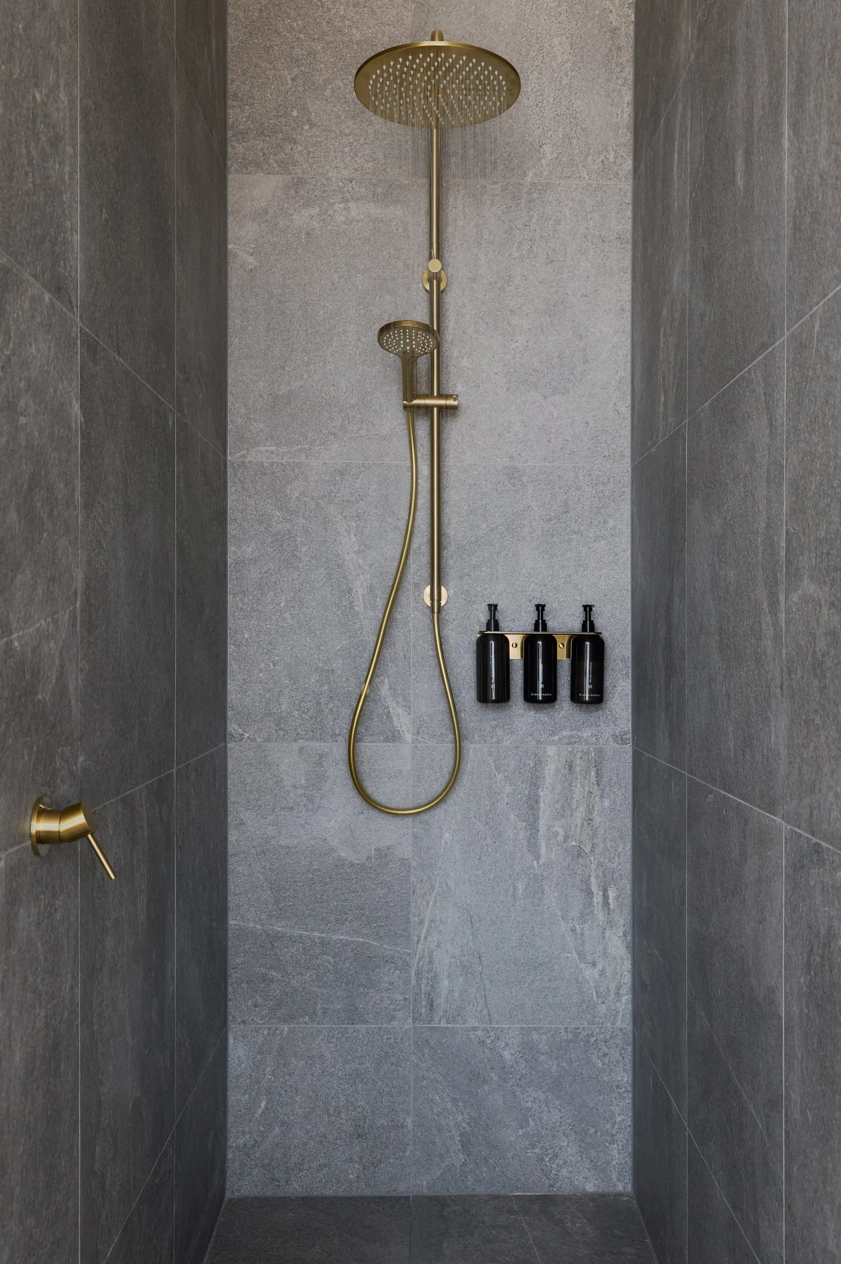 A modern shower area with gray tiled walls and floor, featuring a gold rain showerhead, a separate handheld shower, and three black liquid soap dispensers mounted on the wall.