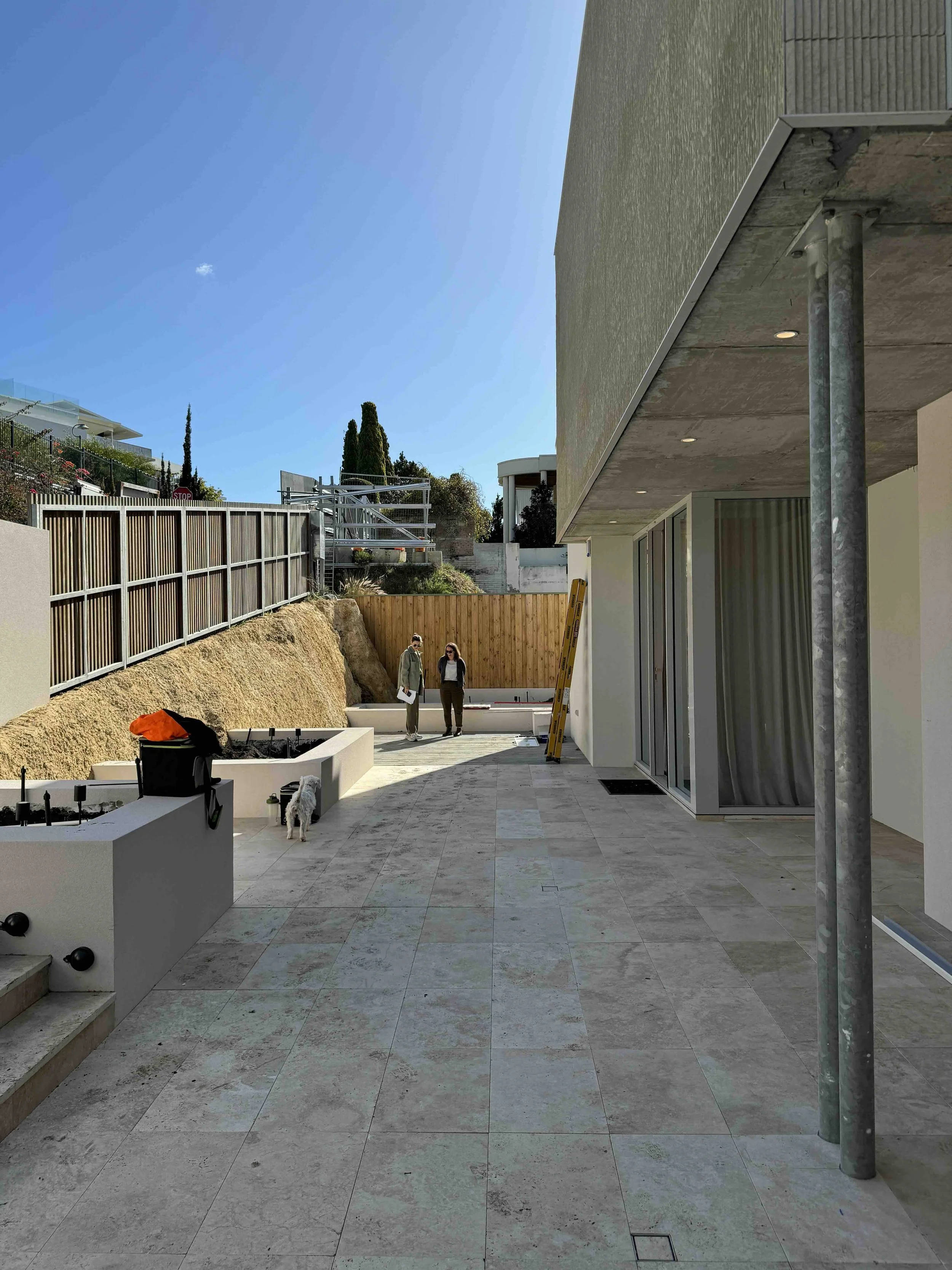 Construction site with two people talking, a dog, and building materials on a partly finished patio area, with a modern house and wooden fencing in the background under a clear blue sky.