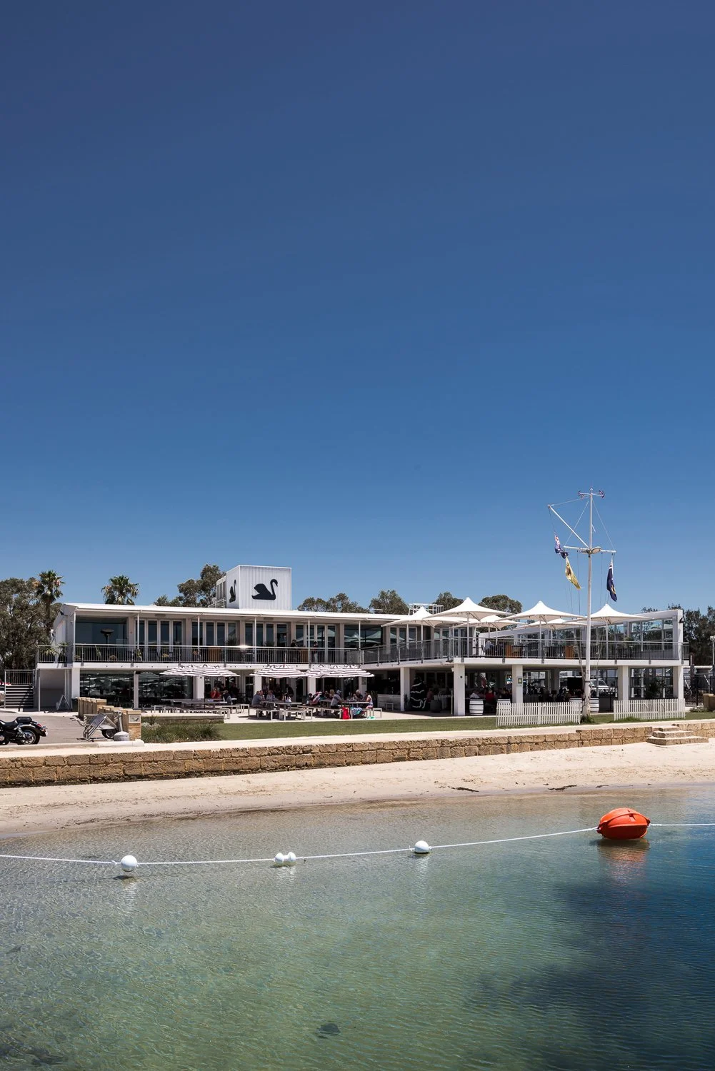 A modern, two-story waterfront restaurant with outdoor seating under umbrellas, and a small boat docked nearby, with a clear blue sky overhead.