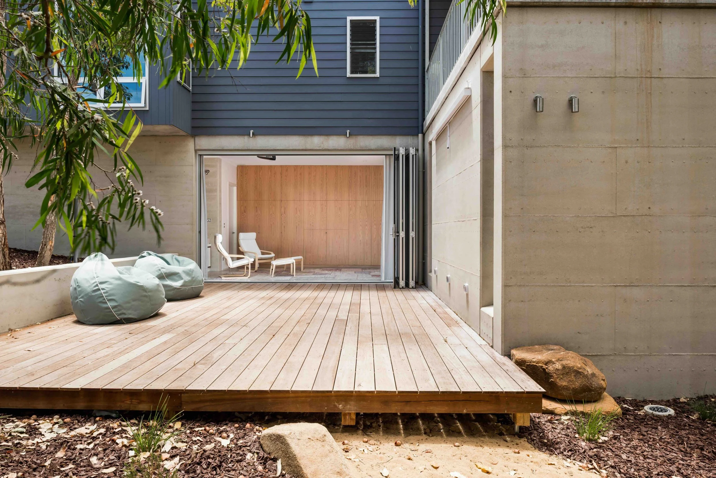 Modern outdoor patio with wooden deck and concrete walls, featuring two green bean bag chairs, a tree, and folding doors opening into a minimalist interior with wooden paneling and white chairs.