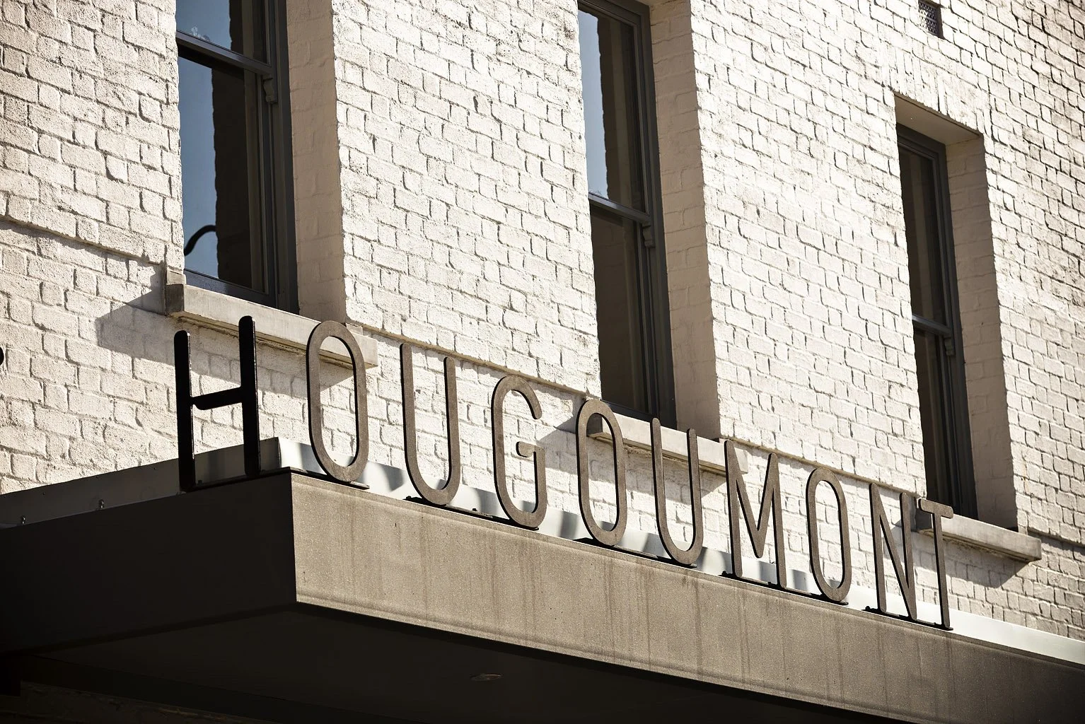 A building with white brick exterior and three windows, featuring a sign that reads 'HOUGOUMONT' in large metal letters.