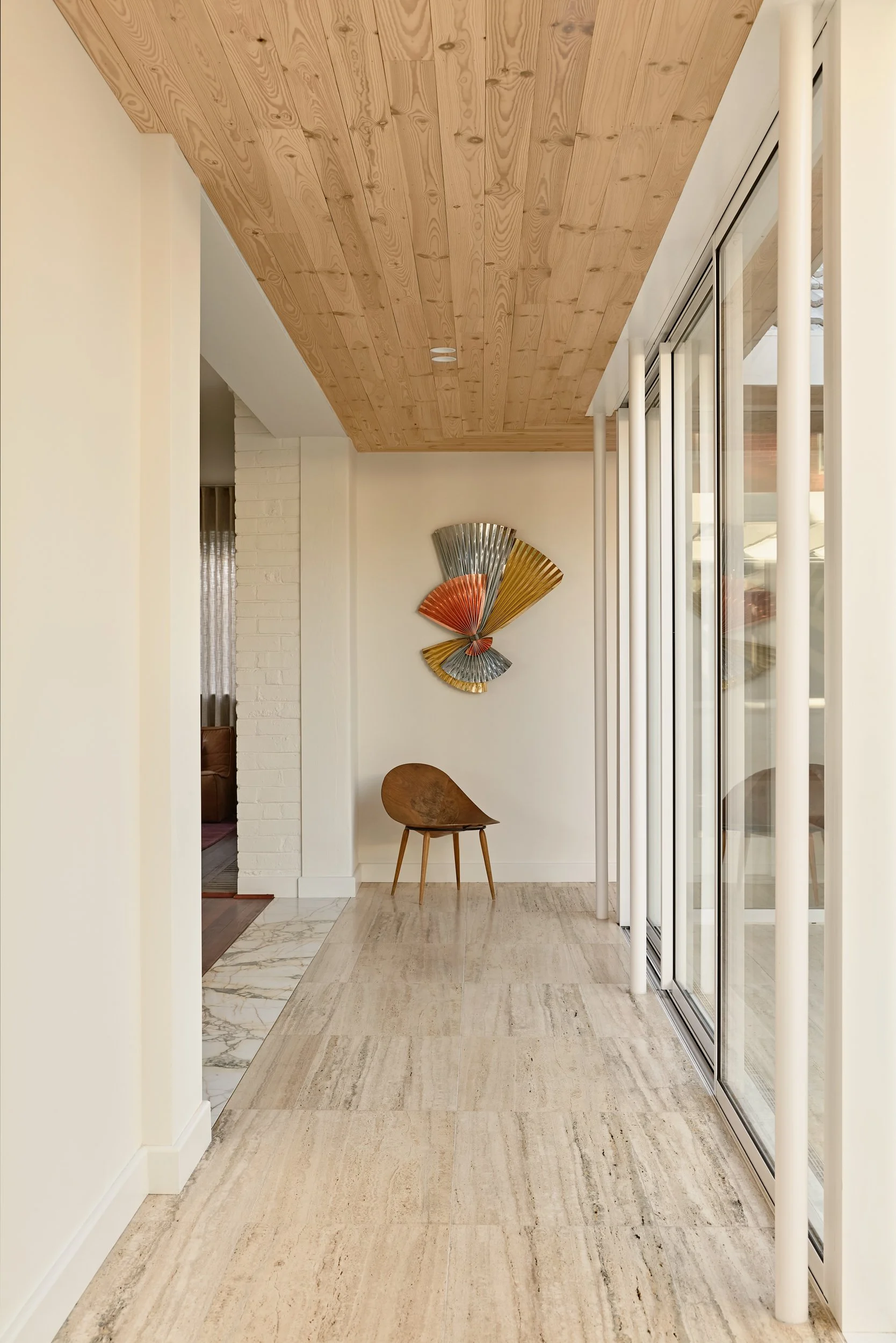 Modern interior space with a wooden ceiling, white walls, a decorative fan art piece, a wooden chair, and large glass sliding doors.