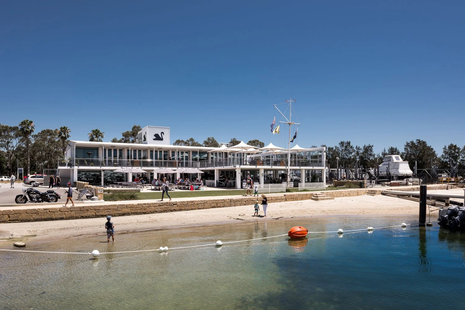 A seaside restaurant with outdoor seating, a boat-shaped sign, and a ship replica, situated by a beach with people walking and kids playing near the water under a clear blue sky.