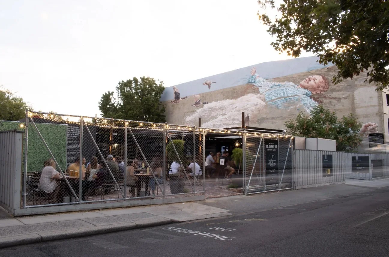 Outdoor restaurant or bar enclosed with chain-link fencing, with patrons sitting at tables under string lights. A mural of a woman reclining and animals is painted on the building wall behind.
