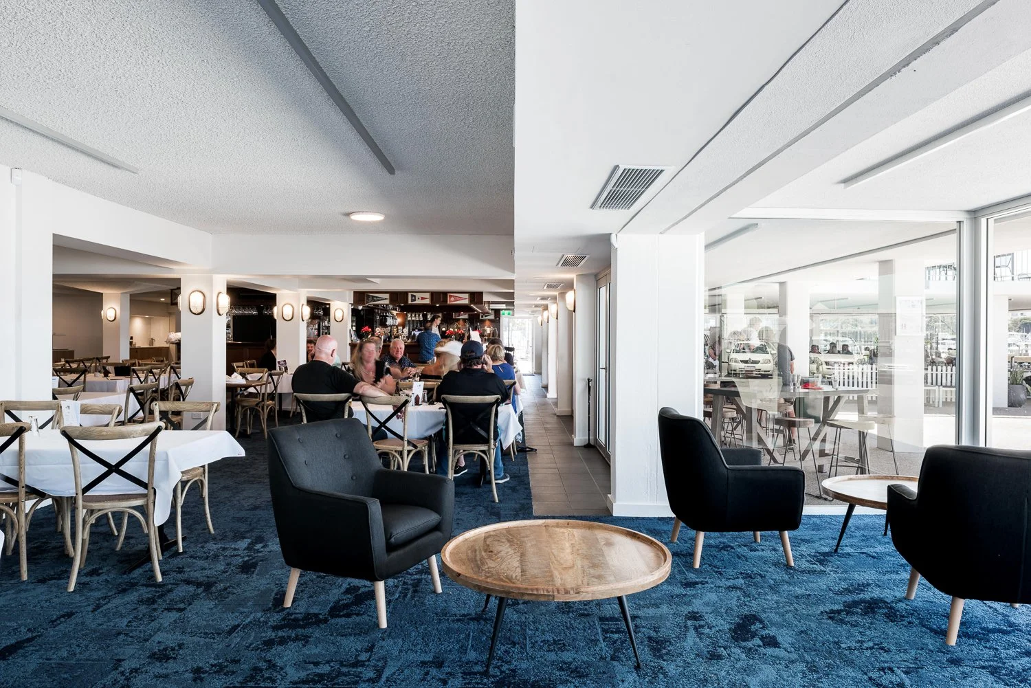 Interior of a restaurant with black armchairs, round wooden tables, and white tablecloth-covered tables with wooden chairs. In the background, people are dining at tables, and large windows let in natural light. Outside, you can see tables, chairs, a