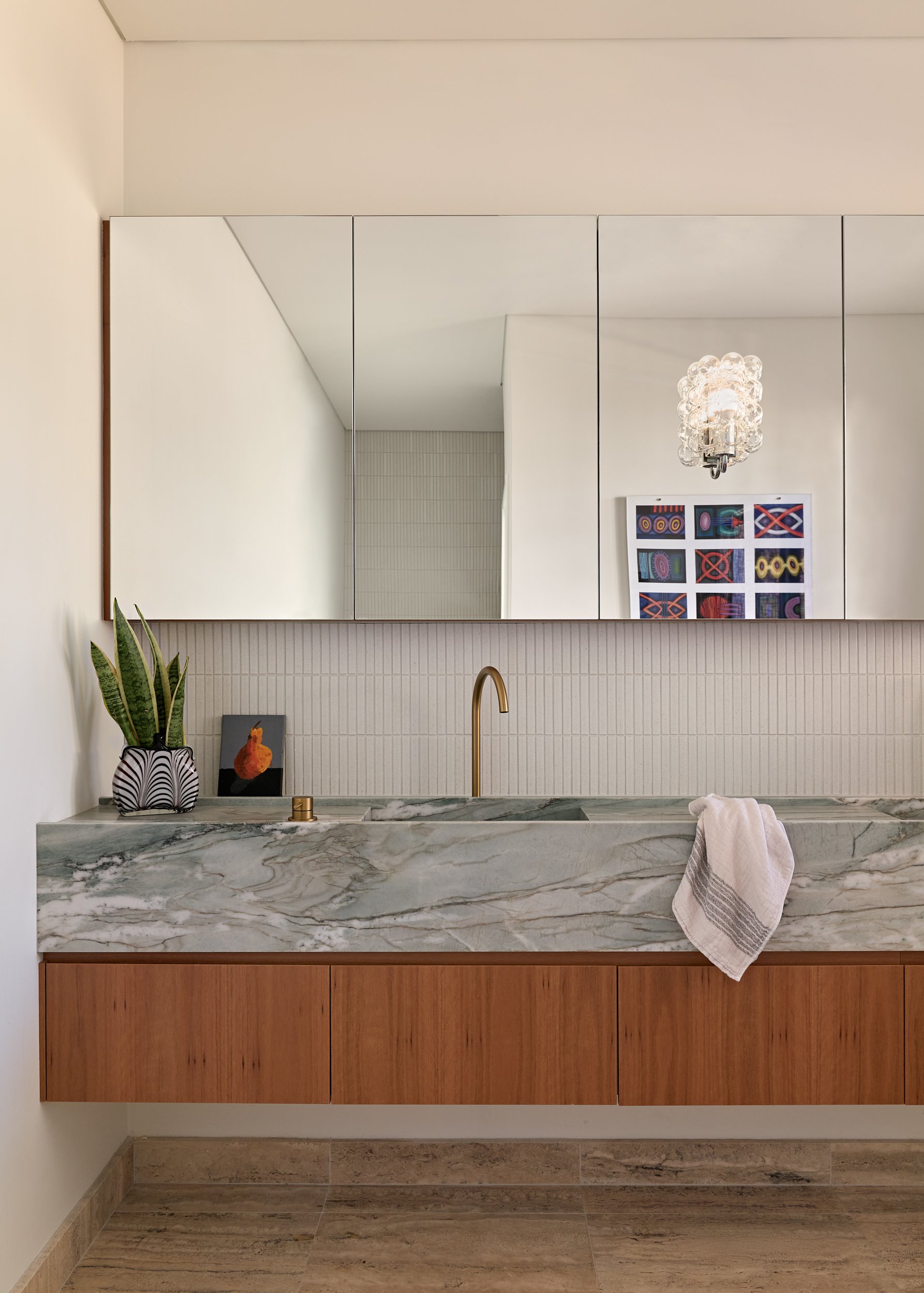 Modern bathroom with large marble vanity, gold faucet, potted plant, and decorative artwork, reflected in a wall mirror with multiple panels.
