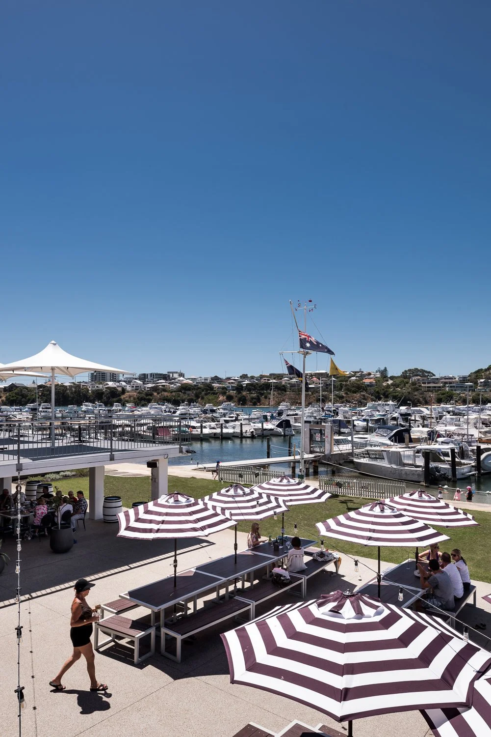 People sitting at outdoor tables with striped umbrellas near a marina with boats and yachts docked, under a clear blue sky.