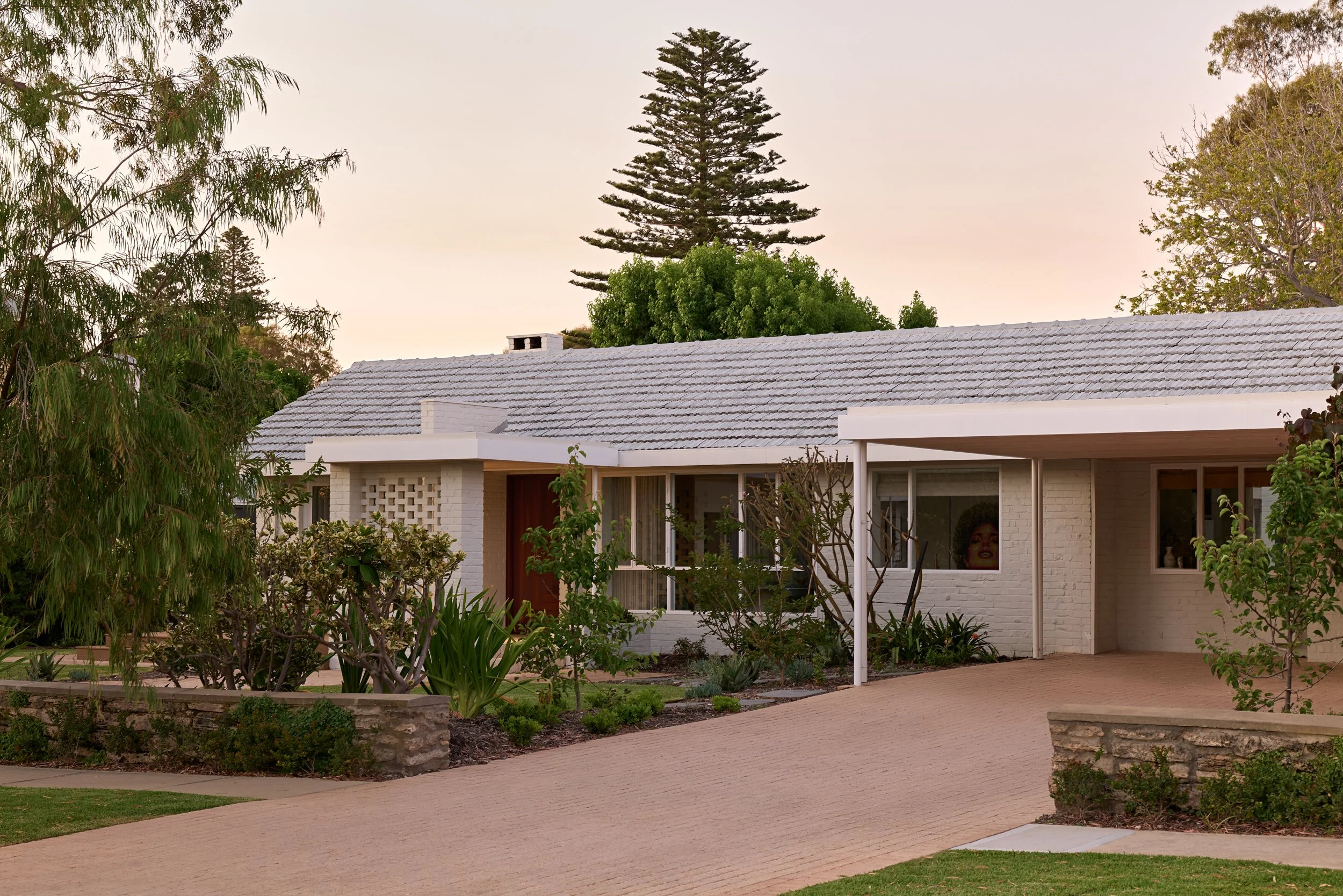 A single-story house with white brick walls, a gray tiled roof, and a small front yard with plants and trees. The house has large windows, and a woman making a face is visible through one of them.